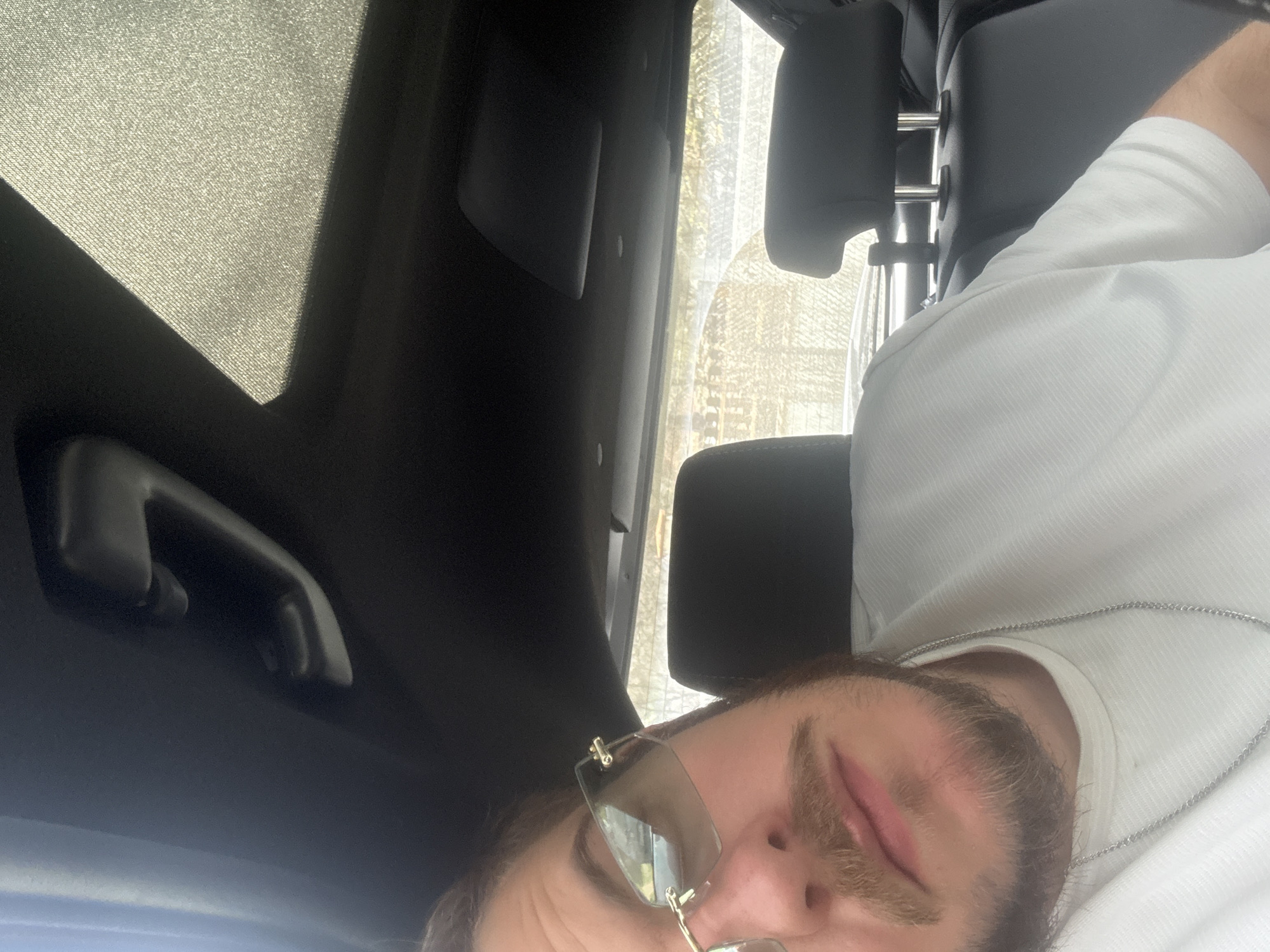 The image shows a man with a beard and sunglasses inside a vehicle, likely a car, due to the visible car interior components such as the grab handle, headliner, and seat. The man is wearing a white shirt and a silver chain. The location is suggested to be Teteven, Bulgaria, but no visible elements confirm this. The man is looking directly into the camera. The lighting indicates daytime. The background through the car window is blurry.