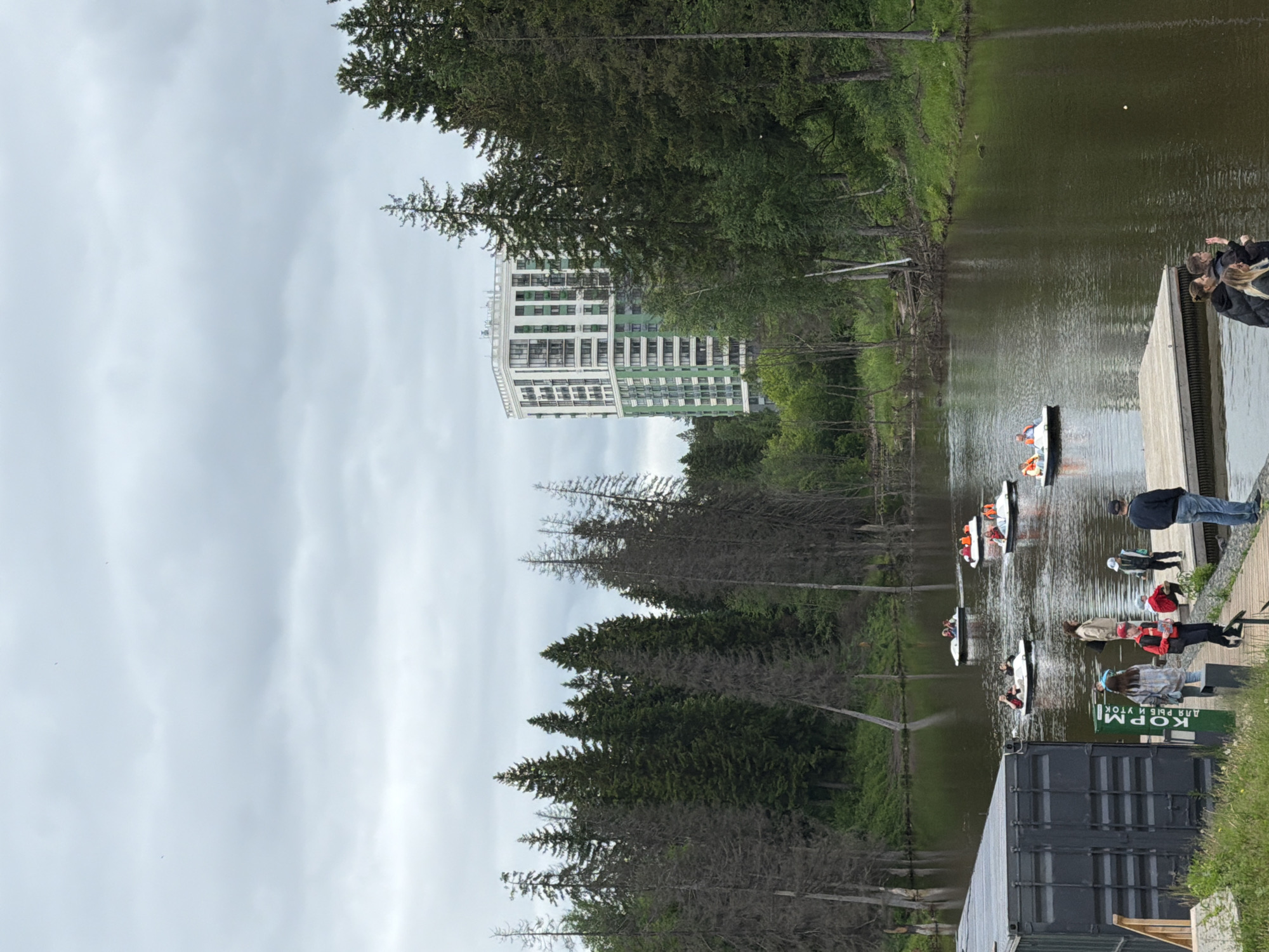 This detailed image captures a recreational scene on a pond in Izhevsk, Udmurt Republic, Russia, specifically near the Park of Culture and Rest named after S.M. Kirov, with the "Vesna" residential complex visible in the background.
The foreground features a dark grey, corrugated shipping container-like structure on the left, acting as a functional building. Adjacent to it, a wooden dock extends into the dark, reflective water. A green sign with white Cyrillic text "КОРМИ ДЛЯ РЫБ И УТОК" (FEED FOR FISH AND DUCKS) stands next to the container. Several people are gathered on and around the dock. From left to right, a person in a light jacket and dark pants stands, followed by a person in a plaid shirt. A person in a red jacket and black pants stands further right. A young person in a white cap crouches on the dock, and a man in a dark jacket and jeans stands at the dock's edge, facing the water. At the far right of the dock, three individuals, including two women and one man, are conversing, with one woman waving.
In the midground, the dark green-brown water of the pond occupies a significant portion of the frame. Six white pedal boats, each occupied by one or two individuals wearing bright orange life vests, are scattered across the water, indicating recreational activity. The water's surface shows reflections of the overcast sky and surrounding trees.
The background is dominated by dense forestation bordering the pond. On the left and center, a mix of tall, dark green coniferous trees and numerous bare or sparsely foliated dead deciduous trees are visible. On the right, the forest appears denser and primarily coniferous. Above the treeline in the central-right background, a tall, modern residential building, identified as the "Vesna" complex, rises prominently. It features a distinctive white facade with interspersed green panels and numerous balconies. The sky above is entirely overcast with a uniform layer of light grey clouds, indicating a cloudy day.