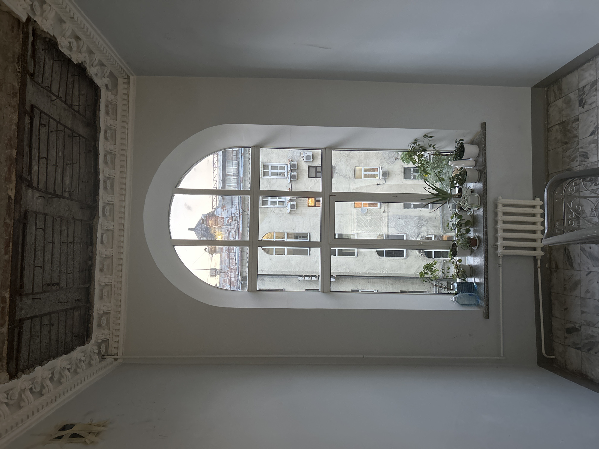 The photograph captures a room with architectural features reminiscent of an older style, possibly in the city of Unknown, Unknown. A large, arched window dominates the composition, offering a view of a neighboring building. The building is characterized by a gray, weathered facade, several windows, and numerous air conditioning units. On the windowsill, a row of potted plants adds a touch of greenery. To the right of the window, there is a white radiator and a section of a marble-tiled wall. On the left side of the photograph, a portion of the ceiling is exposed, revealing a decayed structure with metal support. The overall color scheme is dominated by whites and grays, contributing to a sense of age and neglect.