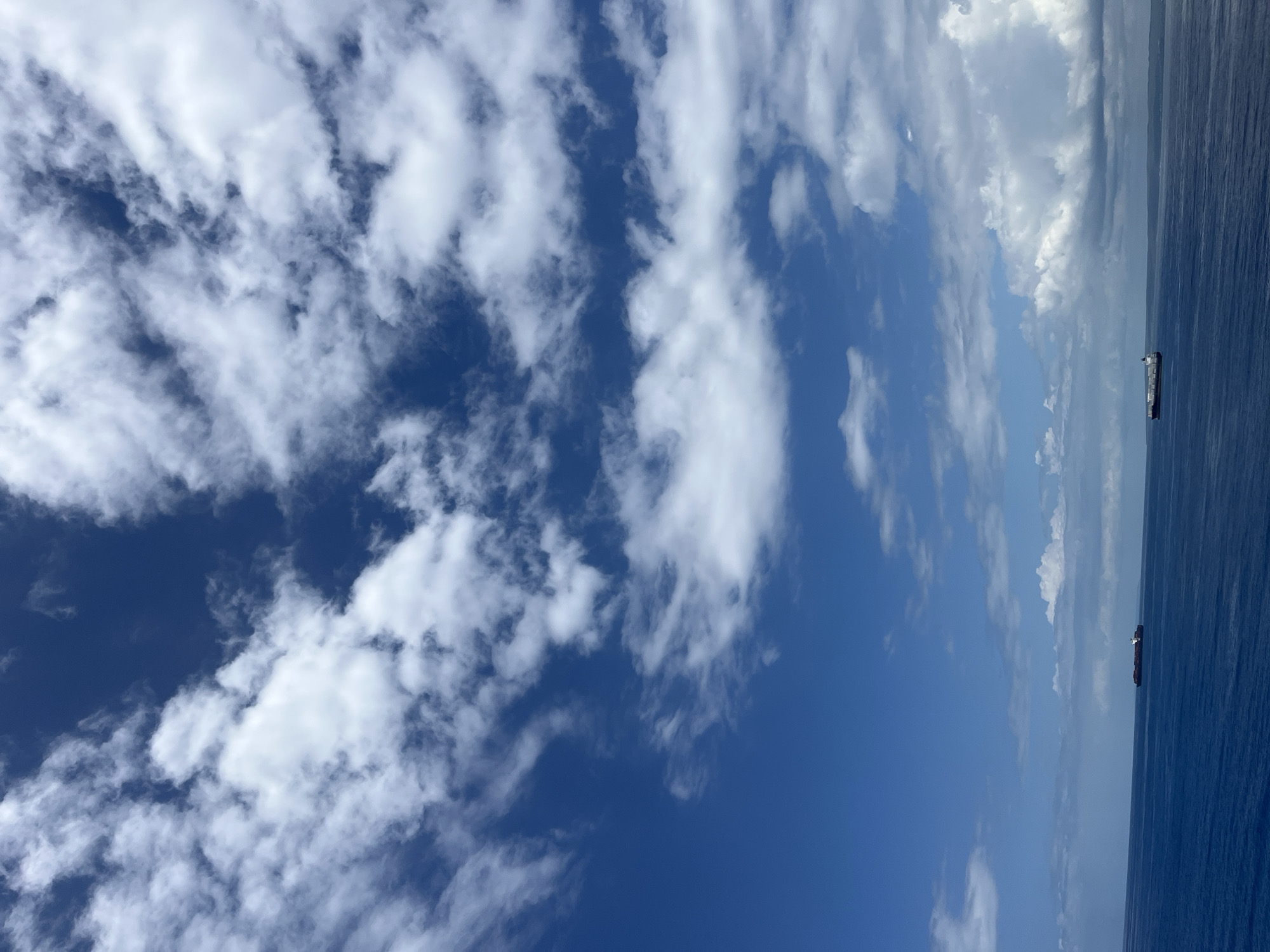 The image presents a view of the sky, primarily filled with white clouds against a blue backdrop. A body of water, possibly the ocean, is visible on the right side of the frame, with two distant cargo ships. The image appears to be taken outdoors during daylight, but the exact location (city, state/province, or country) is Unknown, Unknown.