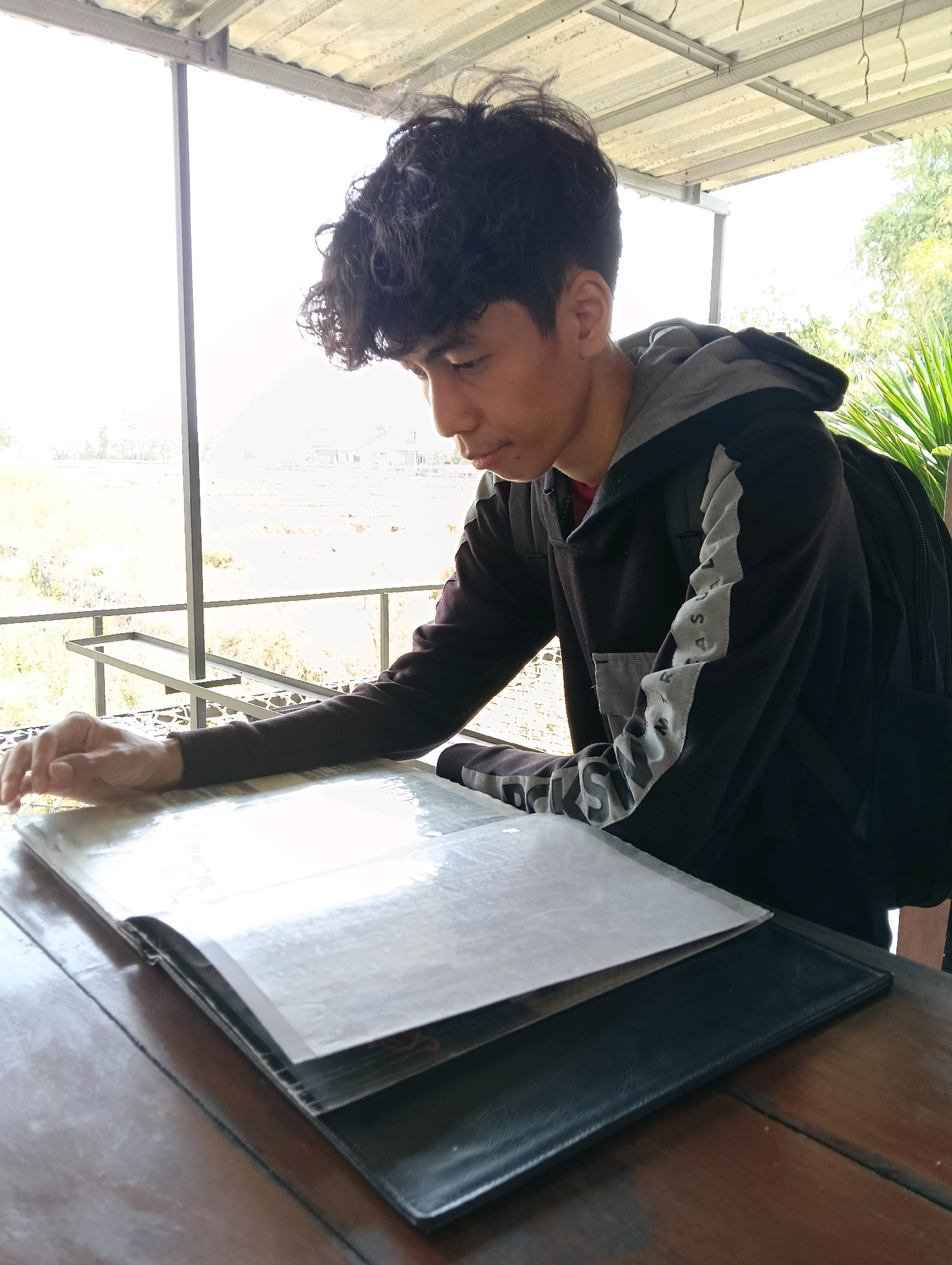 A young man with curly black hair is seated at a wooden table outdoors, looking down at a menu. He is wearing a dark hooded jacket with gray and white accents on the sleeves and a black backpack. His right hand rests on the menu. In the background, there is a bright, hazy outdoor scene with some foliage visible to the right. The setting appears to be a covered outdoor seating area.