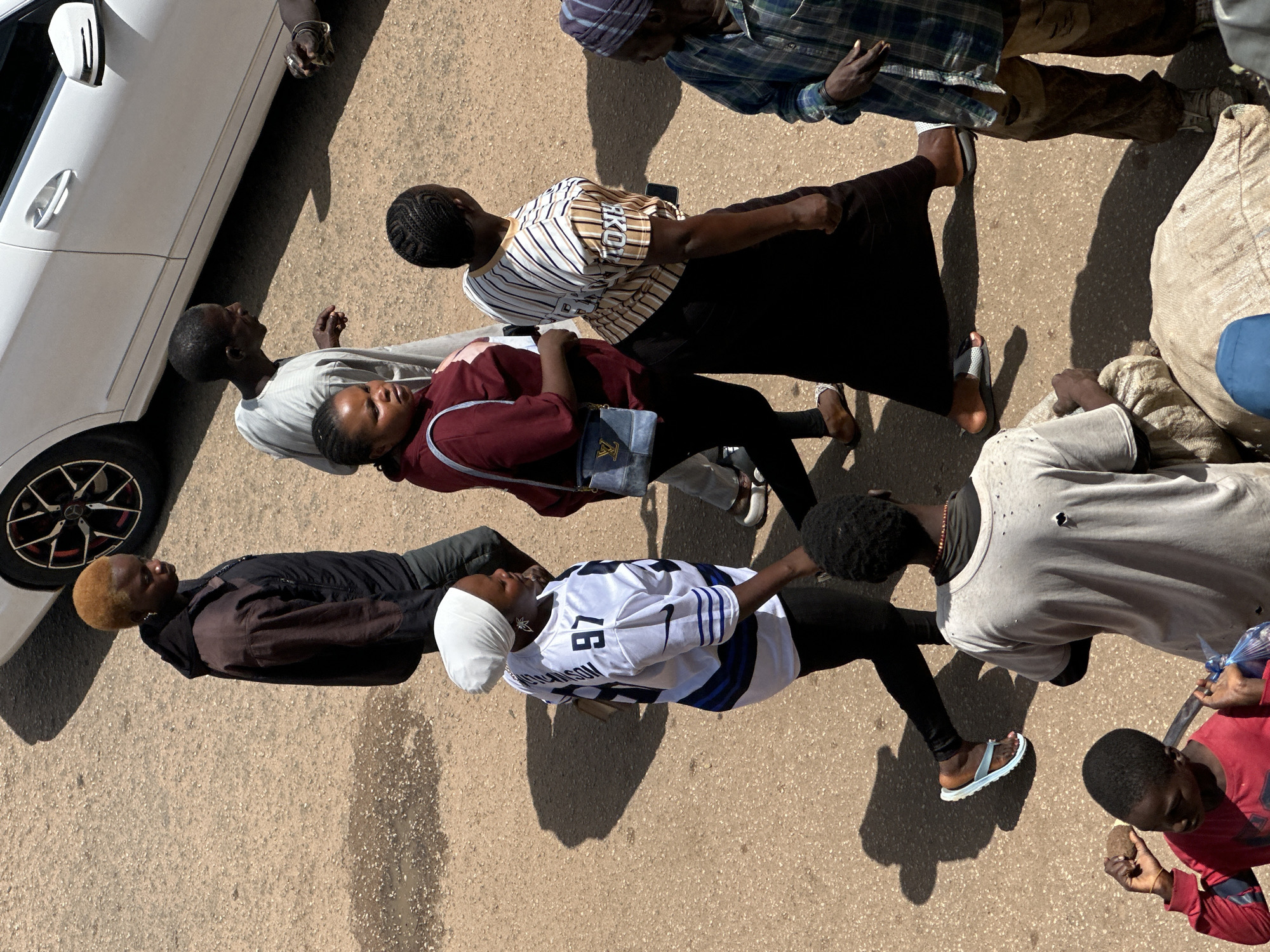 The image shows a group of people on a street in Bukuru, Nigeria. The scene appears to be a casual gathering or a moment of interaction.
The subjects include several individuals, both male and female, of varying ages. One person is wearing a white shirt with blue stripes and the number "97" and "LAKECONN" visible on the back. Another individual is wearing a maroon shirt and a blue bag is slung over their shoulder. A person in the foreground is wearing a brown and black jacket, and another is in a light-colored shirt. There is also someone in a blue and green plaid shirt and another in a white garment.
Objects visible include a white car with a black rim on the wheel. A large burlap sack is also present, along with a blue plastic bag.
The setting is an outdoor street scene, with the ground appearing to be asphalt or a similar paved surface. Shadows are cast on the ground, indicating it is daytime, likely with bright sunlight, suggesting a warm weather day. The overall mood seems neutral, with no overt display of strong emotions. The image is taken from a high angle, looking down on the group.