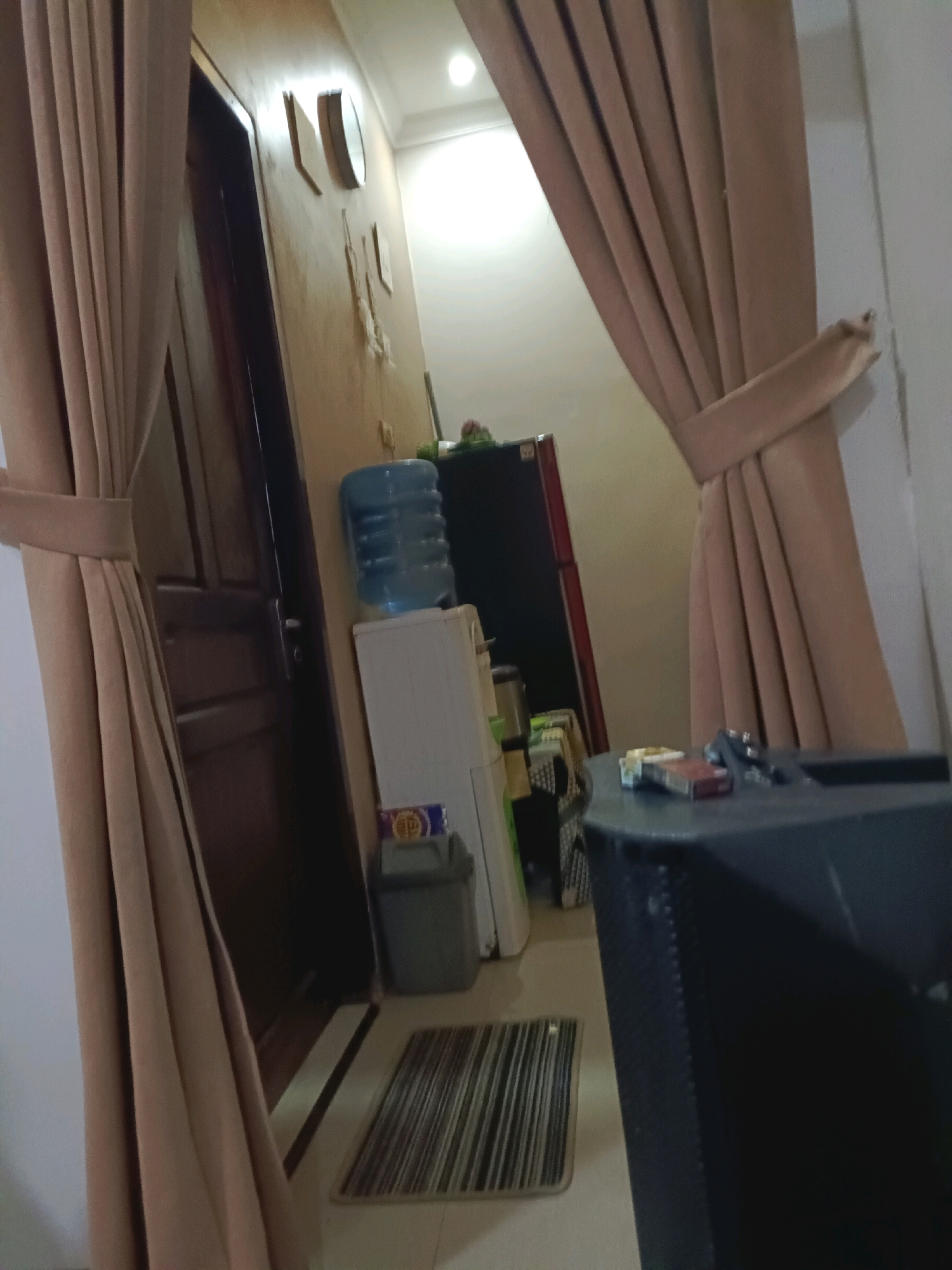 The image shows an indoor scene in Rancang Kencono, Indonesia. In the foreground, a large black speaker box is partially visible on the right. On top of the speaker box, a pack of cigarettes and a lighter are placed. To the left of the speaker box, beige curtains are drawn. Behind the curtains on the left side of the image, a dark wooden door is visible. Further into the room, a white refrigerator is situated next to a blue water dispenser. On the wall above the refrigerator, decorative objects are hung. A small trash bin is placed on the floor next to the refrigerator. A striped doormat lies on the tiled floor in front of the door. The lighting suggests it is daytime.