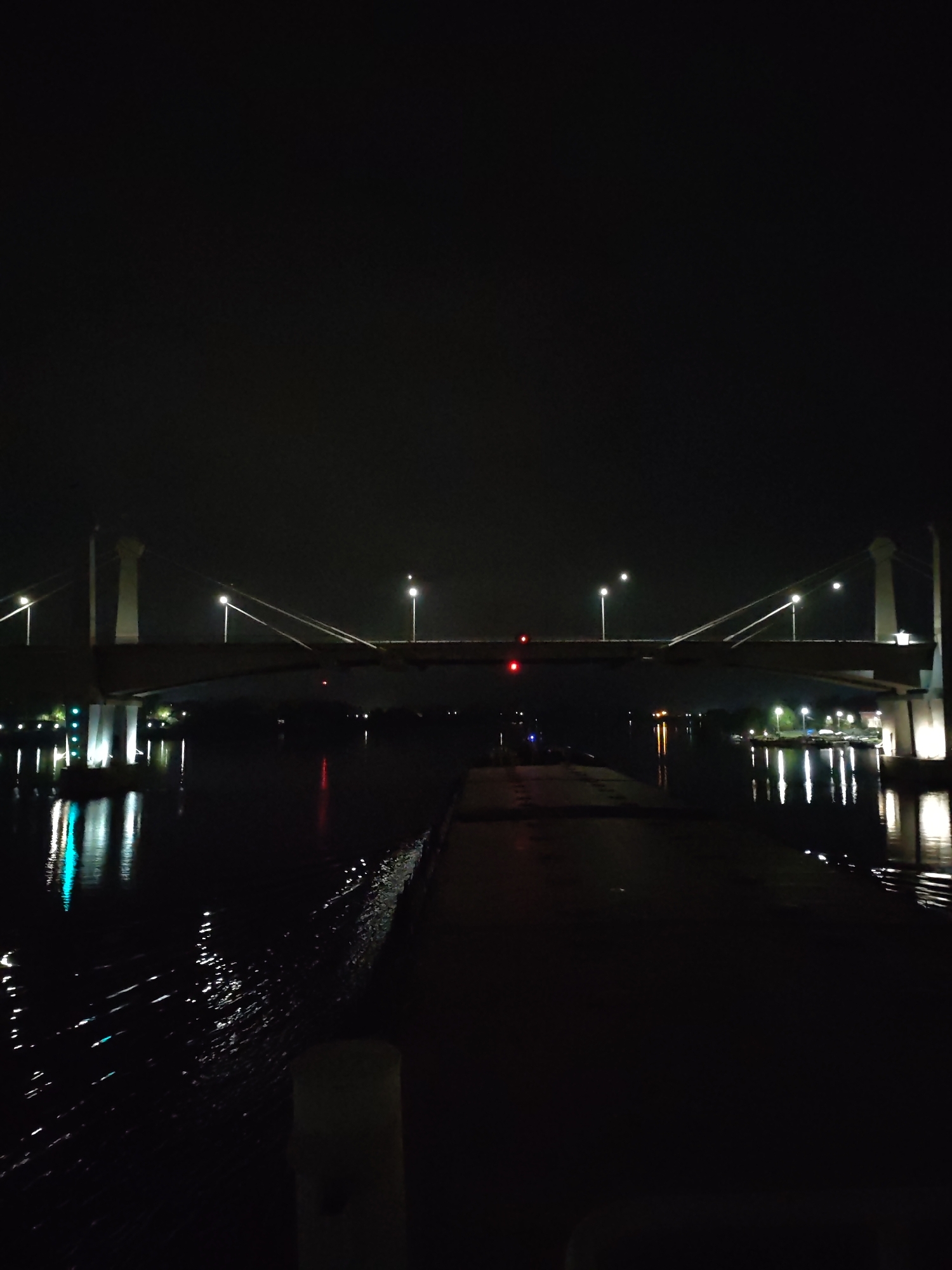 The image shows a night scene in Kimry, Russia, featuring the Kimry Bridge over the Volga River. The bridge is illuminated by several streetlights, which reflect in the water below. There is a dark pier in the foreground leading towards the bridge. A red light is visible on the bridge itself. The sky is dark and cloudy.