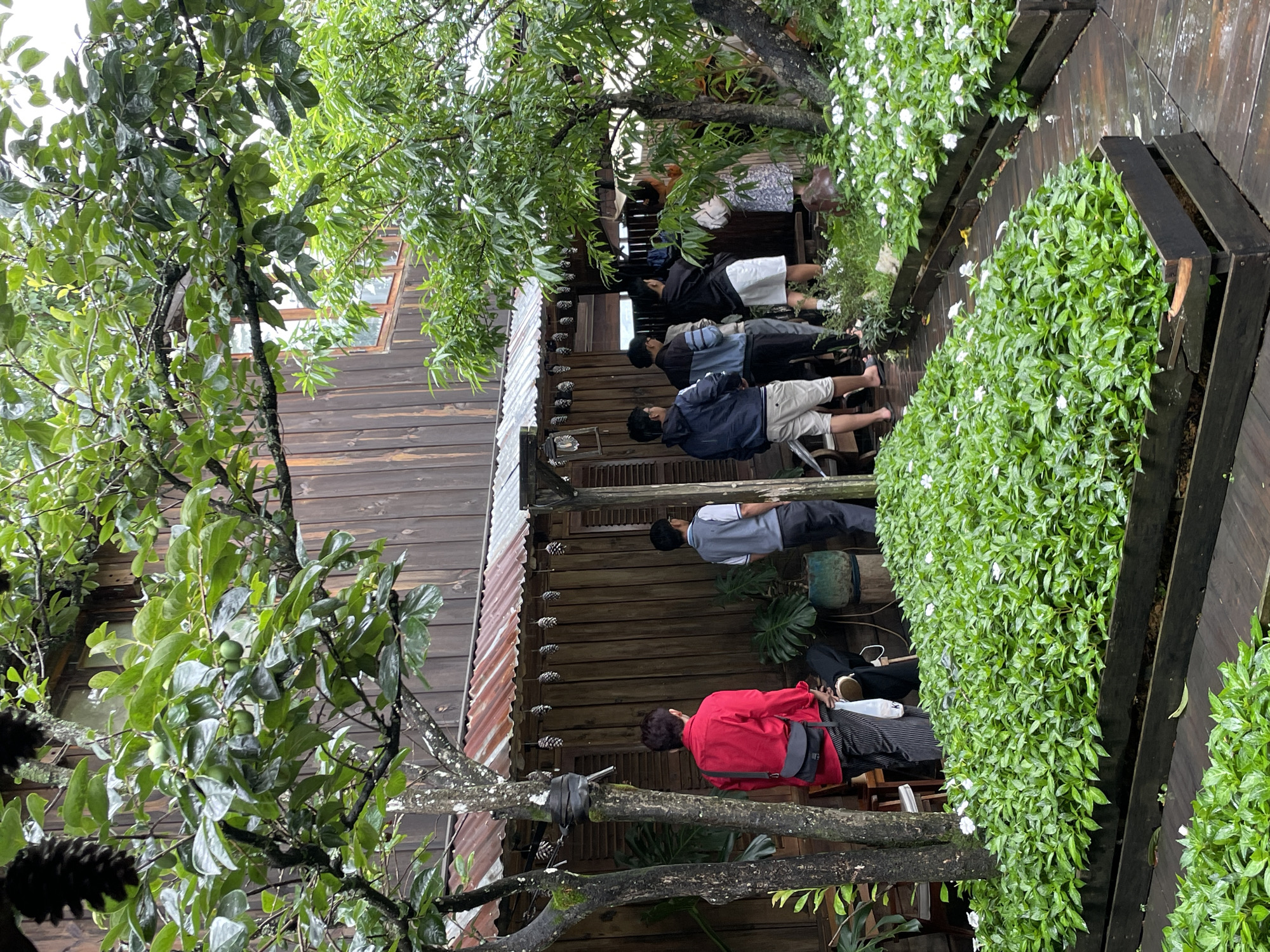 The image depicts a group of people standing outside a rustic wooden building in Da Lat, Vietnam. The building features a corrugated metal roof and wooden shutters. In the foreground, lush green plants with small white flowers are arranged in wooden planters, and a wooden walkway runs between them. Several people are standing near the entrance of the building, some appearing to be queuing. One person in a red shirt is prominent in the left foreground, facing away from the camera. The overall ambiance suggests a natural, possibly cafe or guesthouse setting, with the architecture and greenery characteristic of Da Lat's natural landscape.