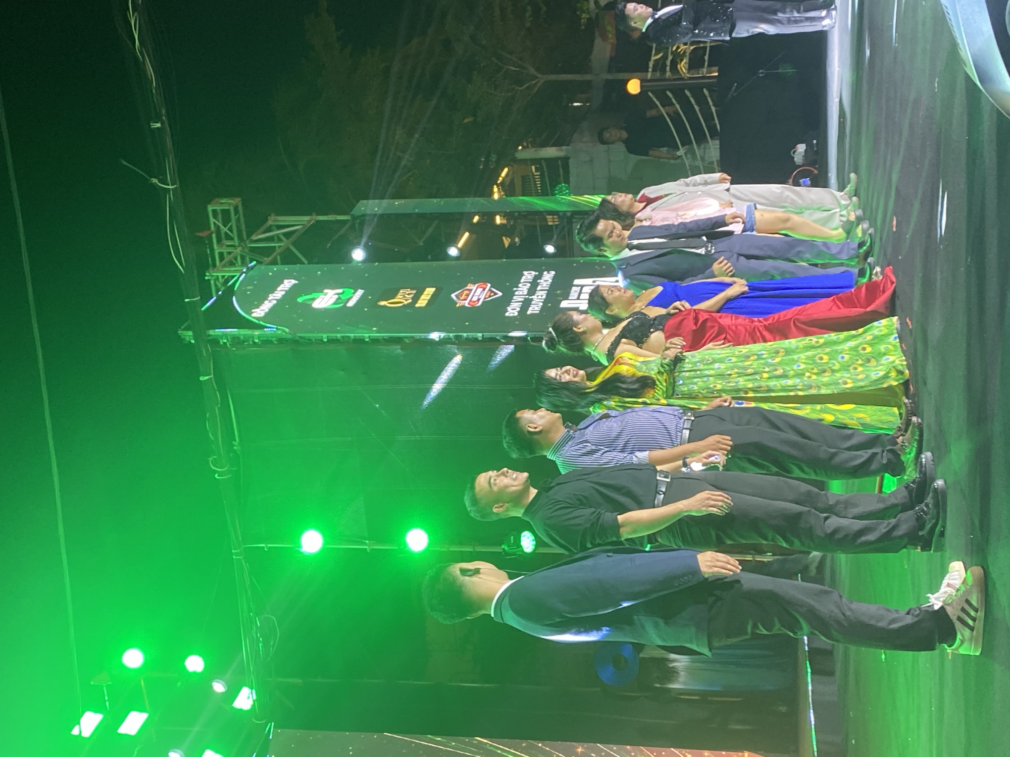The image depicts an outdoor nighttime event, likely a ceremony or performance, taking place on a stage in Mũi Né, Vietnam. A group of people are standing in a line on the stage, illuminated by vibrant green lighting from above. On the left side of the frame, a man in a dark suit stands facing away from the camera, with a man in a black shirt and dark pants next to him, smiling. To his right are several women in colorful dresses, including one in a bright green patterned gown with a high slit, and a woman in a blue dress. Further to the right, men in suits and other attire are also part of the lineup. Behind the individuals on stage, a large vertical banner with logos and text, some in Vietnamese, is visible. The background suggests an open area, possibly near the coast, with trees and structures visible in the darker areas. The overall atmosphere is festive, with the green lighting creating a dramatic effect.