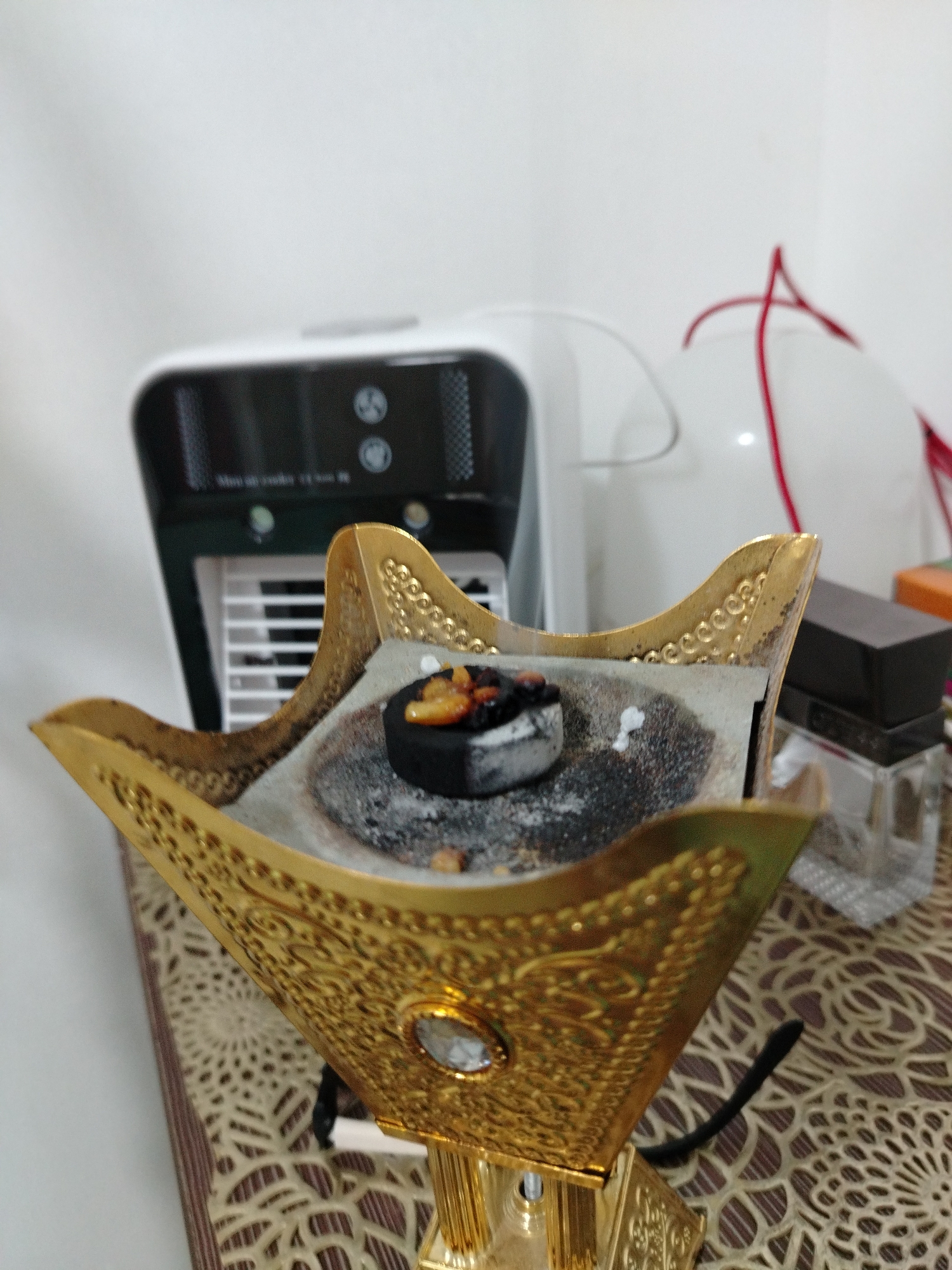 The image shows an ornate, golden incense burner, with a black charcoal briquette smoldering on top, leaving ash and small pieces of frankincense. The burner sits on a table covered with a patterned tablecloth. In the background, there is a white portable air cooler, a white balloon with red cords, and a small square bottle with a black cap. The specific location is not discernible from the image. The image contains no people, interactions, or actions beyond the burning of incense. The environment appears to be indoors.
