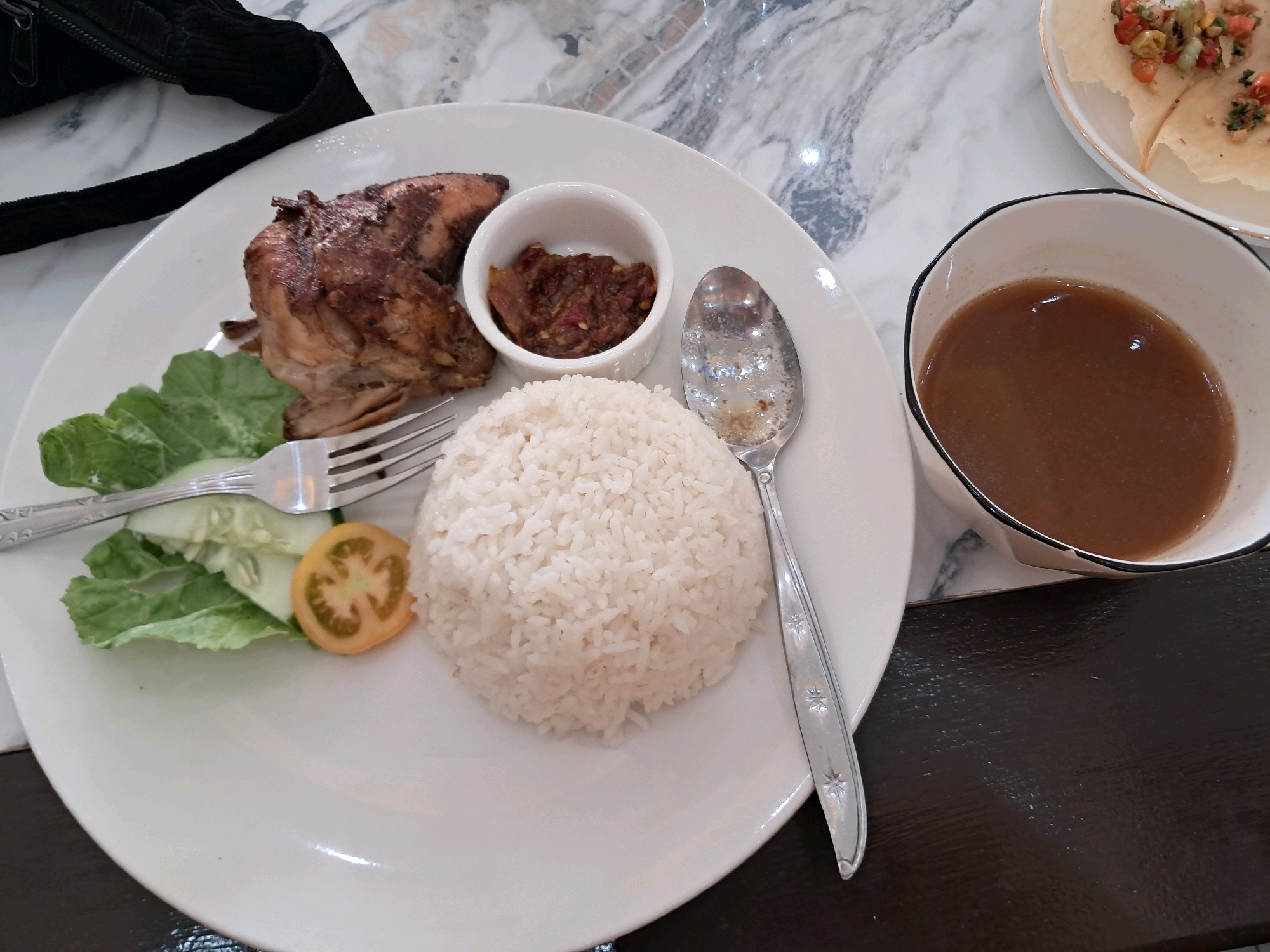 The image shows a close-up view of a plate of food served on a marble-patterned table, likely in a restaurant in Mempawah, Indonesia. The plate contains a portion of roasted chicken, a small bowl of sambal (chili paste), a mound of white rice, and a garnish of lettuce, cucumber slices, and a tomato slice. A fork and a spoon are also on the plate. To the right of the plate is a bowl of brown sauce, possibly a gravy or broth. In the background, a black bag or purse is partially visible. The lighting suggests an indoor setting.