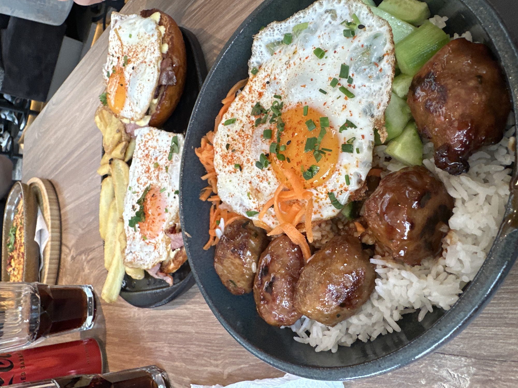 The image depicts two plates of food, presumably at a restaurant or cafe. The food is primarily Asian-inspired, and is located in Sofia, Bulgaria.
Plate 1 contains a rice bowl with a fried egg on top, shredded carrots, sliced cucumbers, and several glazed meatballs. The rice is white and fluffy. The egg yolk is orange. The meatballs appear to be made of pork or beef.
Plate 2 contains a dish with fries and some sort of open-faced sandwich. The fries are pale yellow. The sandwich has a slice of bacon, an egg, and something beneath it.
The table is made of wood and there are some glasses and a bottle of liquid on it.