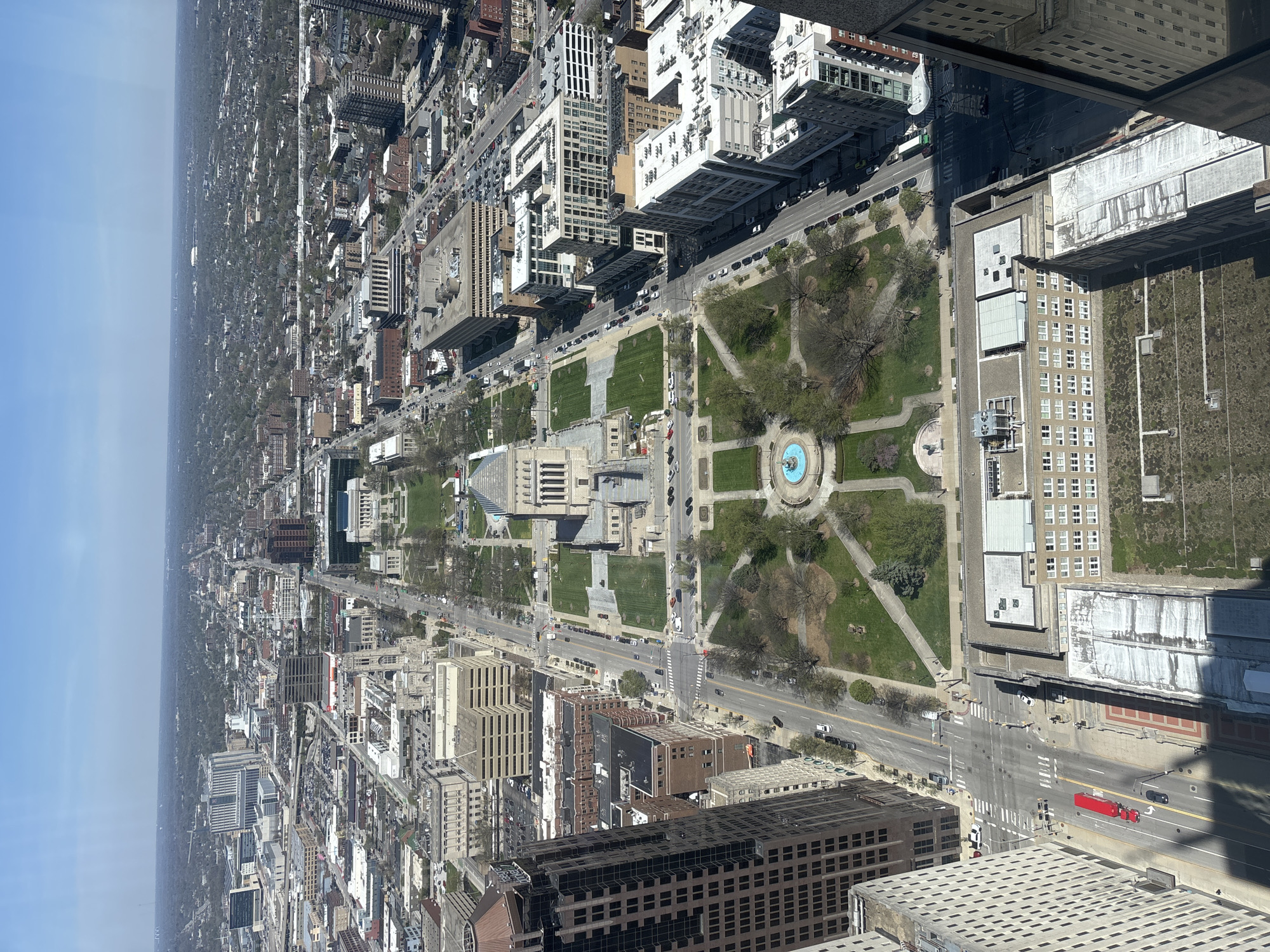 This image shows a high-angle, rotated aerial view of downtown Indianapolis, Indiana, United States. The Indiana Statehouse is visible in the center of the frame. Monument Circle, with a central fountain, is adjacent to the Statehouse. The image includes streets with vehicles, buildings of varying heights, parks, and trees. The sky appears bright and clear.