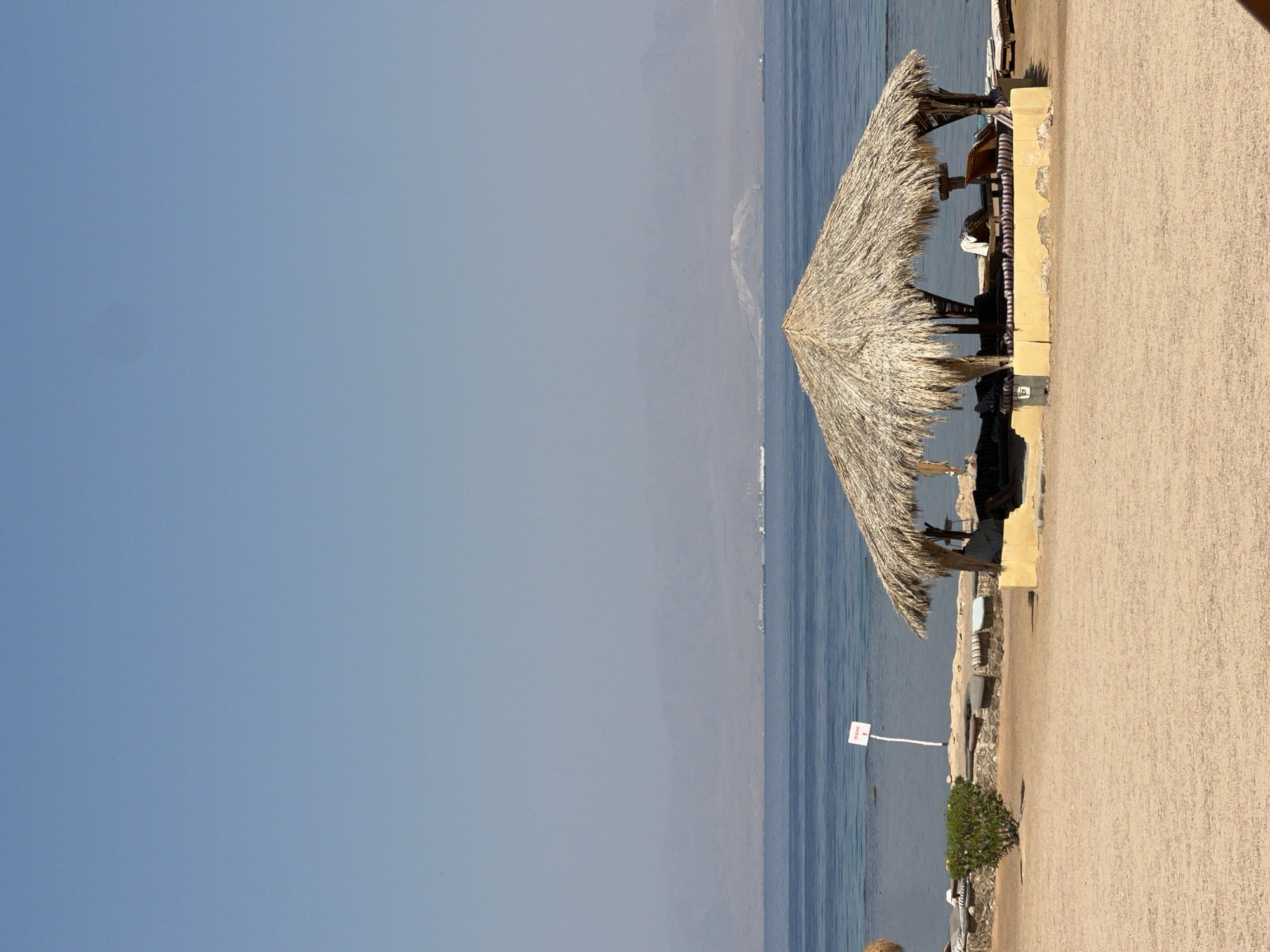The image depicts a beach scene in Nueiba, Egypt, on a clear day. In the foreground, sandy beach stretches towards the water. A thatched-roof gazebo with yellow-painted base sits on the sand, offering shade. Inside the gazebo, lounge chairs are visible. To the left of the gazebo, a small green bush stands out against the sand. A white sign with red text on a pole is planted in the sand near the water's edge. The sea is calm and blue, with a hazy mountainous horizon in the distance. Ships are visible on the water. The sky is a pale blue.