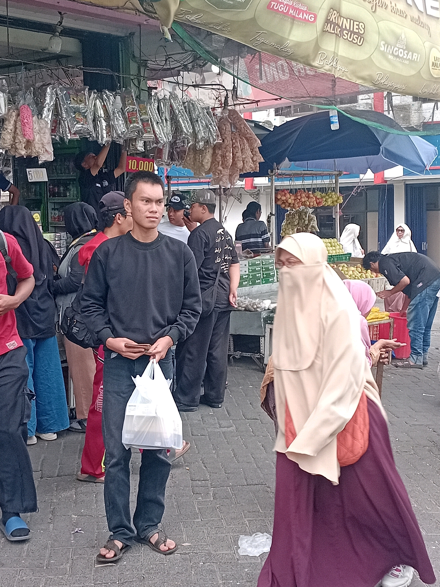 A street scene in Lawang, Indonesia, features a bustling market with various vendors and shoppers. The foreground is dominated by a man wearing a dark sweatshirt and jeans, holding a plastic bag and a wallet. To his right, a woman in a light beige hijab and a dark purple dress is walking away from the camera, with an orange bag slung over her shoulder. In the background, several other individuals are visible, some browsing at stalls and others walking through the market. Stalls are laden with goods, including packaged snacks hanging from above, fruits, and other items. Umbrellas and tarpaulins are set up to provide shade. The ground is paved with stone blocks.
