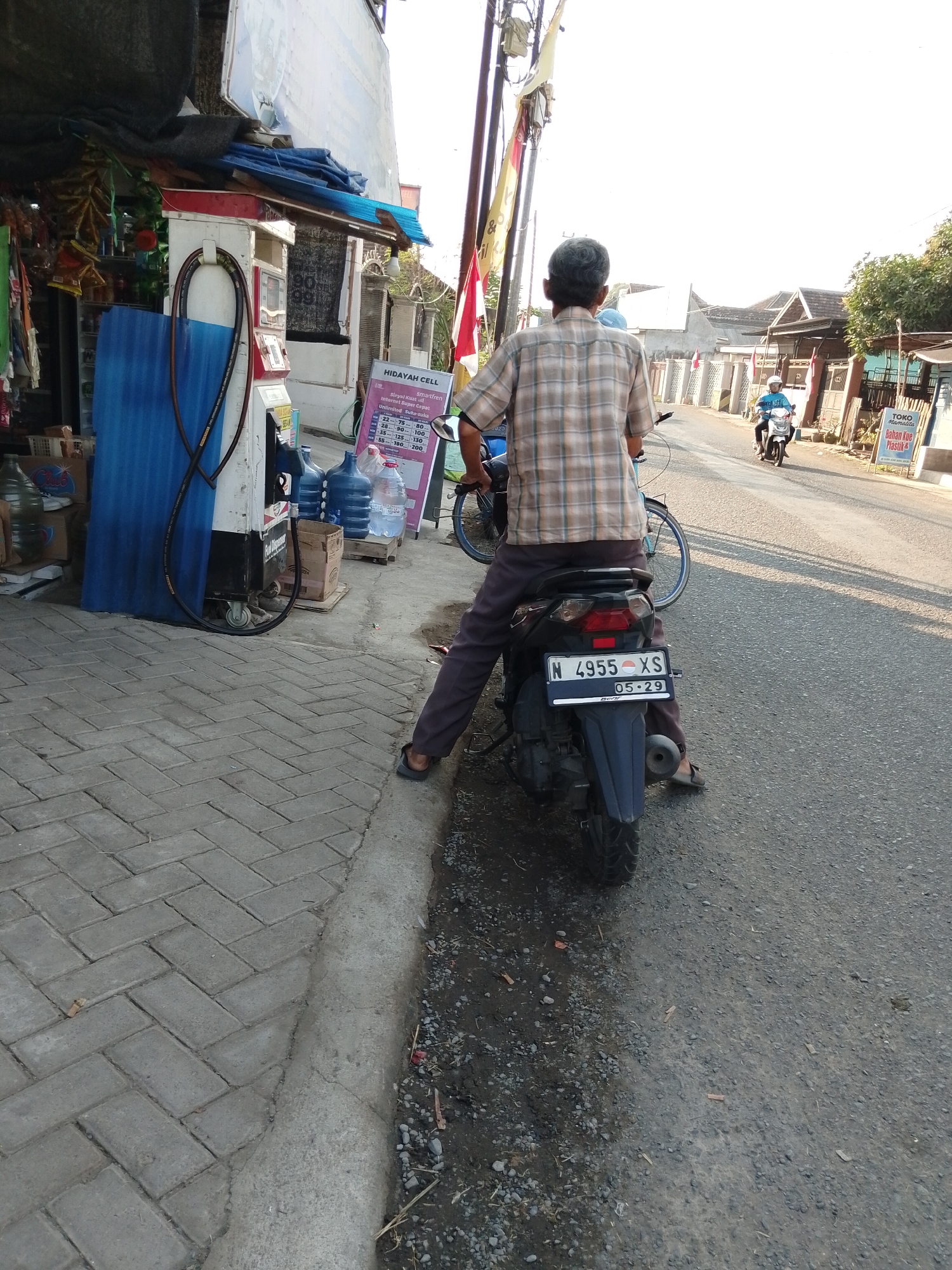 A man in a plaid shirt and dark pants sits on a black motorcycle on the side of a paved road in Pasuruan, Indonesia. The motorcycle has a license plate that reads "N 4955 XS" and a date sticker showing "05-29". To the left of the motorcycle is a red and white gasoline pump. Behind the pump, there are several large water jugs and a display of items for sale. The man is facing away from the camera, looking down the street. In the distance, another person is riding a white motorcycle. The scene appears to be a street in a residential or commercial area, with buildings and trees visible in the background. The sun is setting, casting long shadows.