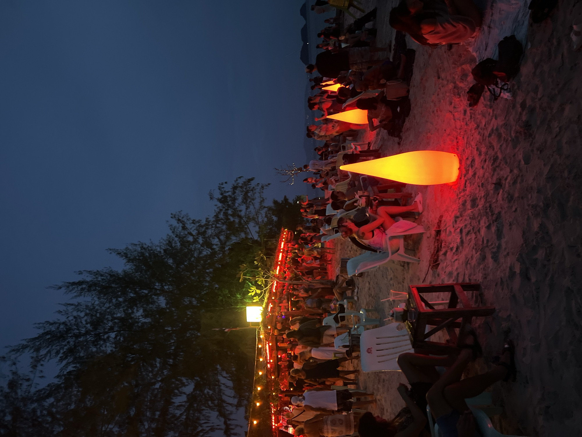 A night-time scene unfolds on a sandy beach in Ko Pha-ngan District, Thailand, illuminated by the warm glow of lights. A bustling beach bar, adorned with numerous red string lights, serves as the focal point on the left side of the image. A diverse crowd of people, many of them socializing and standing, are gathered around the bar. Several individuals are seated on chairs and mats scattered across the sand, some engaged in conversation while others appear to be relaxing. Prominently featured on the beach are three large, cone-shaped illuminated lamps, two of which are brightly lit in orange and red hues, casting a vibrant ambiance. Pine trees frame the scene on the left, their dark silhouettes contrasting with the illuminated sky and the lively atmosphere. The ocean is visible in the background, with faint outlines of islands in the distance, suggesting a coastal setting. The ground is covered in sand, with a few small tables and chairs interspersed among the people.