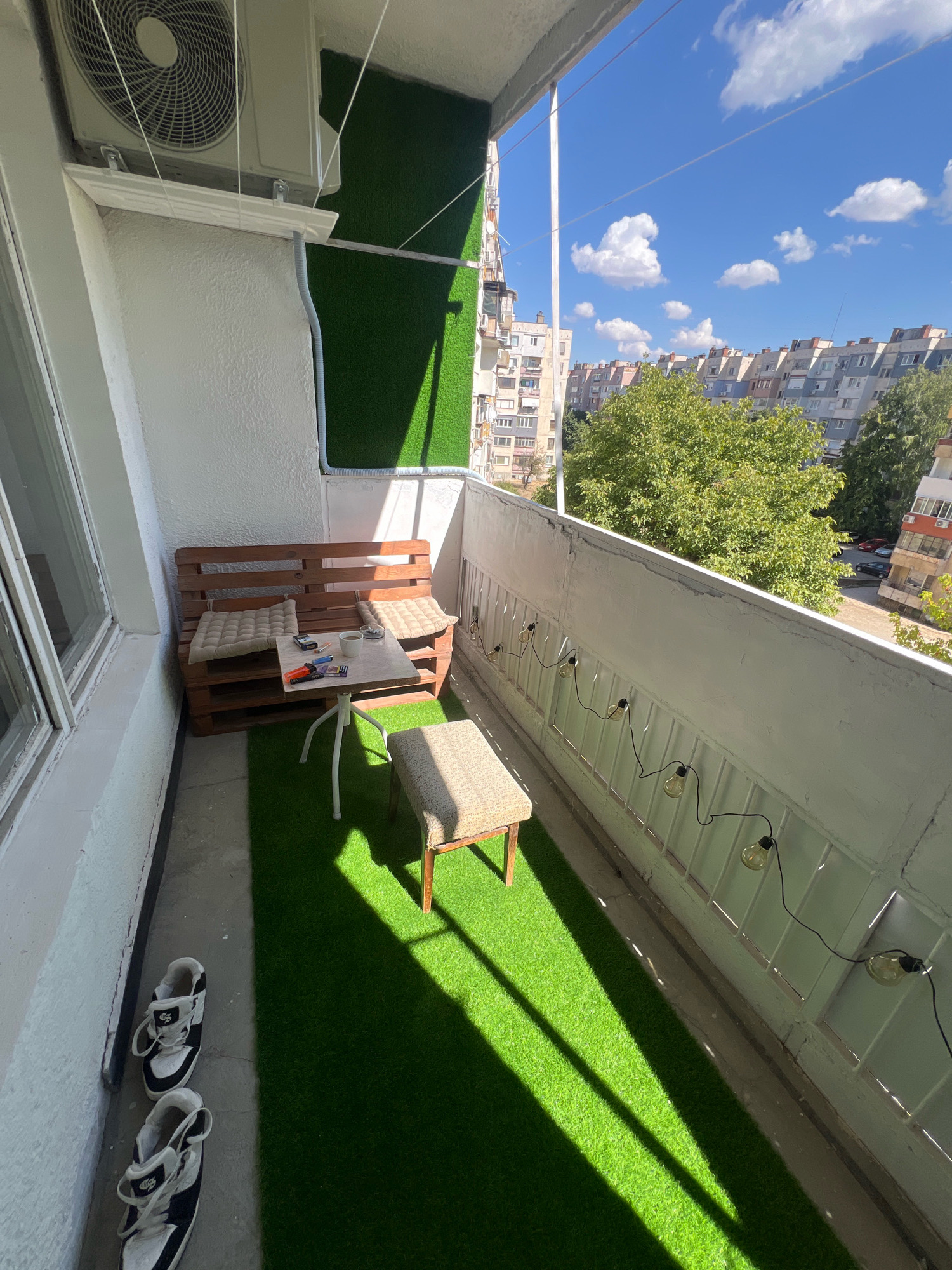 This image depicts a balcony in an urban setting, possibly in Montana, Bulgaria, as suggested by the apartment buildings visible in the background. The balcony is covered with artificial turf, creating a vibrant green surface.
In the foreground, a pair of black and white athletic shoes are placed on the concrete floor near a white wall.
The balcony features a small seating area. A bench made from wooden pallets is adorned with two beige cushions. In front of the bench, a small, light-colored table with a white metal base holds a few items, including a cup of coffee, some cigarette packs, and what appear to be lighters. Next to the table, a small, upholstered stool with a patterned fabric complements the seating arrangement.
The balcony railing is white and features a string of decorative lights with exposed bulbs.
An air conditioning unit is mounted on the wall above the seating area. The surrounding environment outside the balcony shows typical residential apartment buildings with multiple floors and windows. Green trees are also visible, suggesting a mild climate. The sky is bright blue with scattered white clouds, indicating a sunny day. Sunlight casts long shadows across the artificial turf, highlighting the time of day.