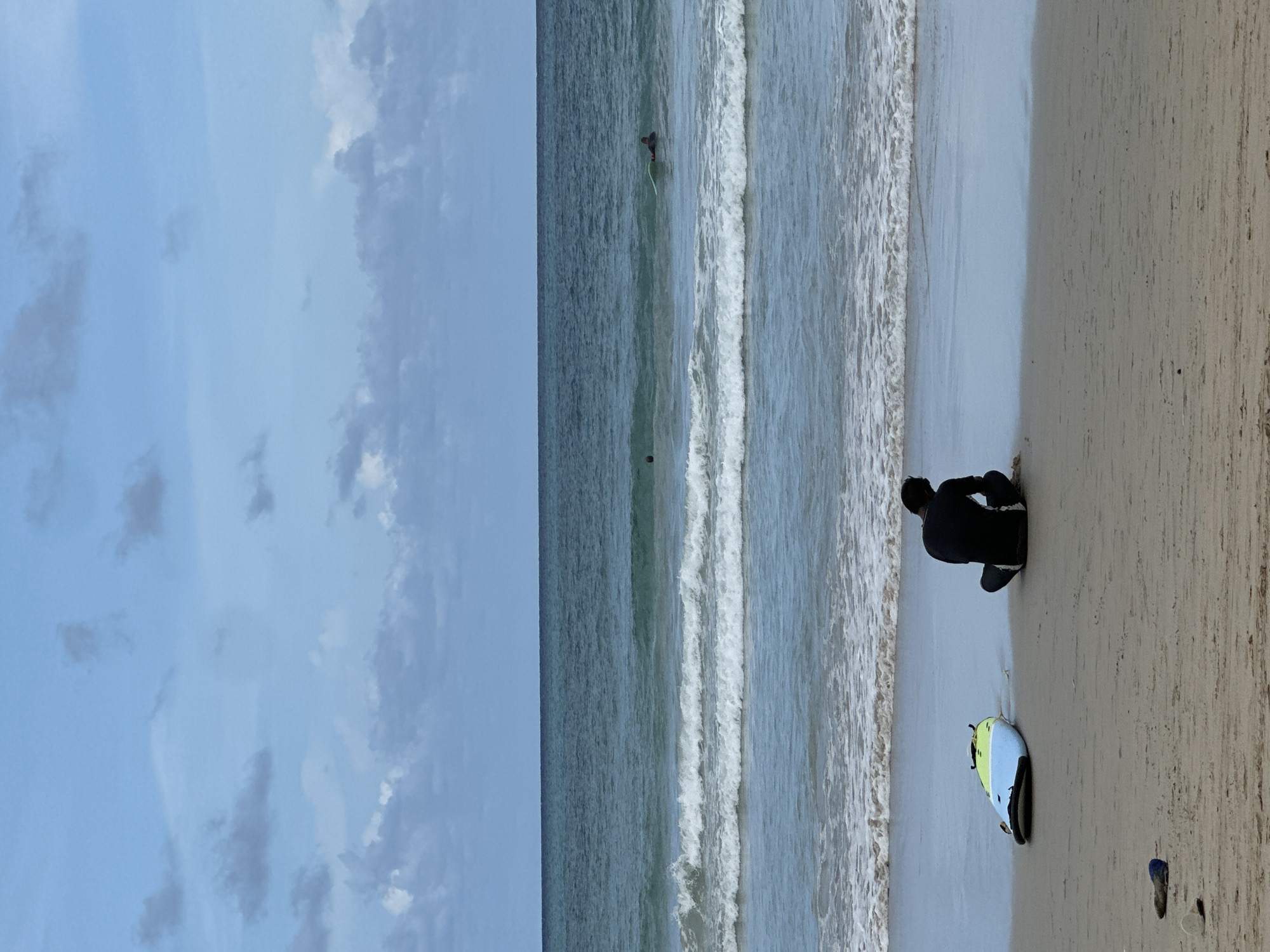 In Karon, Thailand, on a beach with sand and gentle waves, a person in a black wetsuit sits facing the ocean, their back to the viewer. To the left, a surfboard with blue, yellow, and white accents rests on the sand. In the distance, two surfers are visible in the water, one closer to the shore on a light-colored board, and another further out. The sky is overcast with a mix of blue and gray clouds, and the ocean is a deep blue-green.