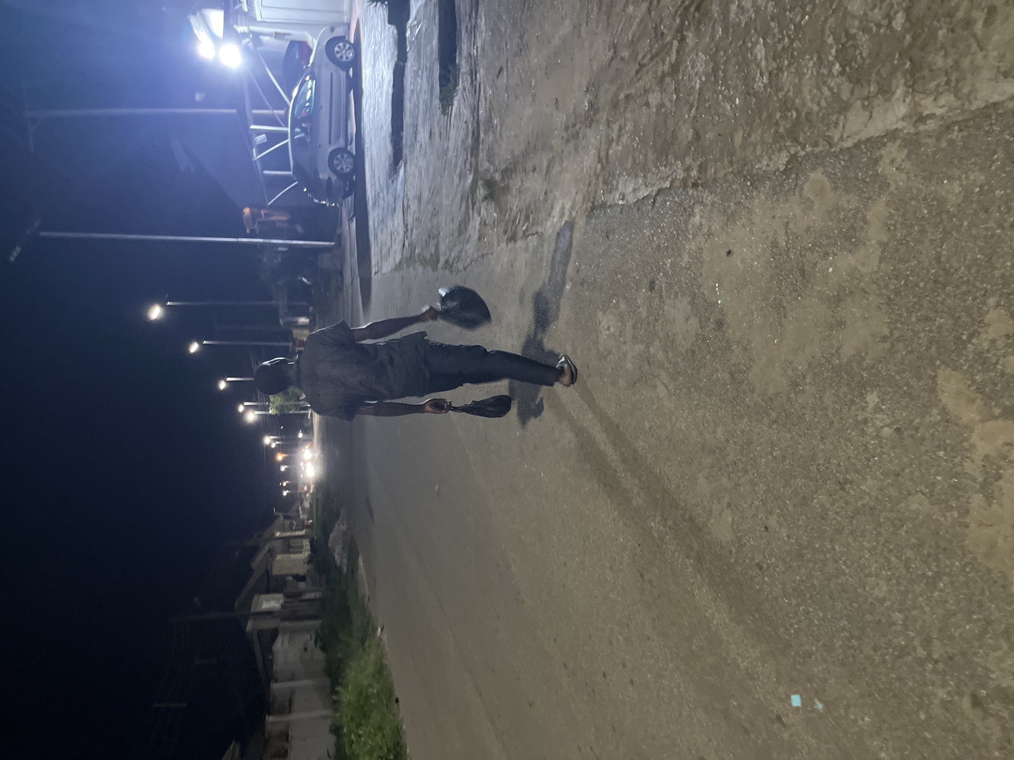 A person walks away from the camera on a paved road at night. The person is wearing a dark t-shirt and dark pants, with light-colored flip-flops. They are carrying a black bag in their left hand and a black helmet in their right hand. The road is illuminated by streetlights and the headlights of distant vehicles. Houses and fences line the left side of the road, and a silver car is parked on the right side. The environment appears to be a residential street in Abuja, Nigeria, likely a suburban or less developed area. The ground is wet in places, suggesting recent rain.
