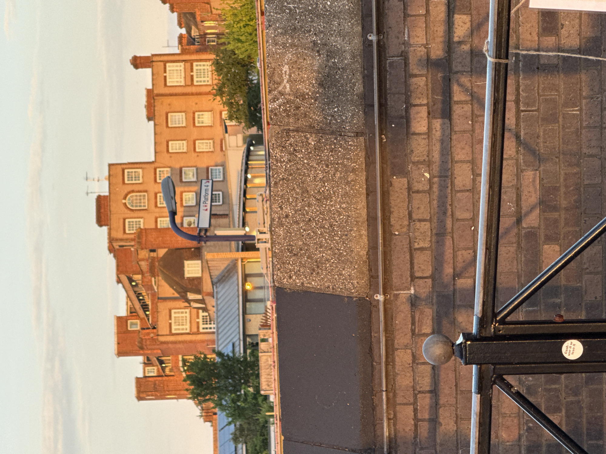 Here's a description of the image:
The image is a street-level view in what appears to be a residential area in the city of London, United Kingdom. A brick building with multiple windows is visible in the background, along with a blue and white platform sign. The building is constructed of orange-toned brick. In the foreground, a portion of a concrete wall or barrier is seen, followed by a brick-paved area. The edge of a black metal railing is visible in the right foreground. A small round sticker with text on it is attached to the railing post. Some greenery is also visible, with trees partially obscuring the buildings behind the concrete barrier. The time of day appears to be late afternoon or early evening based on the lighting.