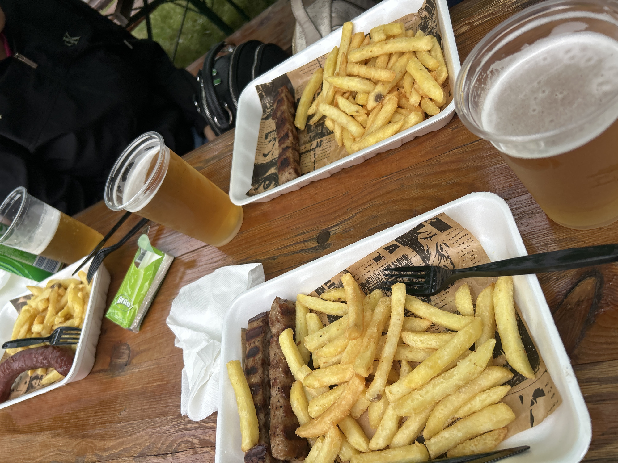 An overhead, eye-level medium shot captures a close-up of two white rectangular takeout containers filled with french fries and meat, as well as two plastic cups of light-colored beer, all resting on a wooden table. The container in the foreground on the left holds a portion of french fries and three grilled meat skewers. A black plastic fork is resting on the fries. To the right of the first container, a second white takeout container is visible, containing french fries and two grilled meat skewers. To the right of this, a clear plastic cup of beer is partially in frame. A second clear plastic cup of beer, with a foamy head, is in the bottom right corner of the image. In the background, a person wearing a black jacket is partially visible, and a dark bag is resting on the table. The overall scene suggests an outdoor dining setting.