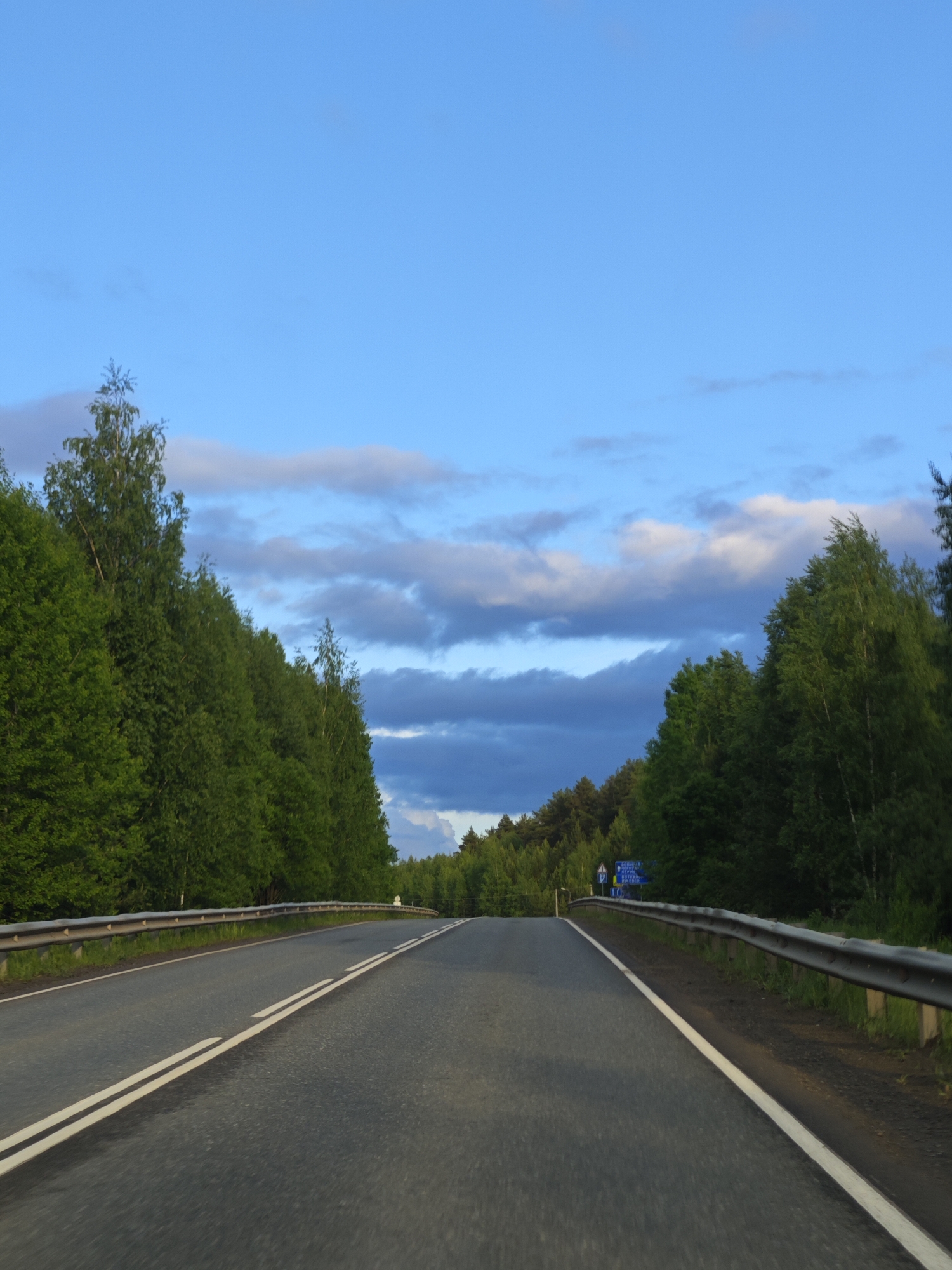 The image depicts an asphalt road stretching into the distance, flanked by dense green trees on both sides under a partly cloudy blue sky. The road is marked with double white lines along its center. A metal guardrail runs alongside the road's edge. A sign is visible on the right side of the road, although its text is illegible. The photograph appears to be taken at ground level, suggesting the perspective of someone driving. This is an outdoor setting on the outskirts of a town or city given the absence of buildings and abundance of trees. The location is somewhere in Chaykovsky, Russia.