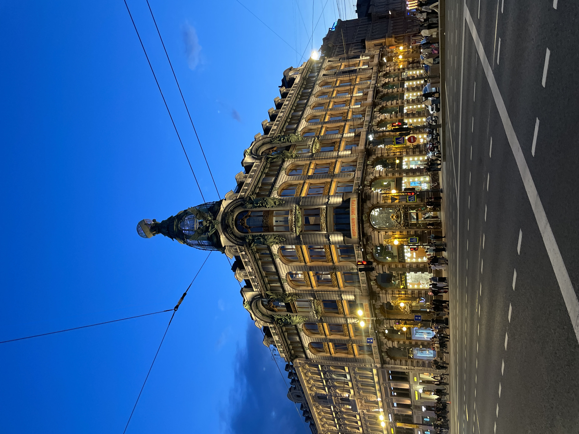 The image displays the iconic House of Books (Dom Knigi) building in Saint Petersburg, Russia, captured during dusk or early night. The large, ornate building, originally constructed for the Singer Company, dominates the left side of the frame, its many windows and intricate facade details illuminated with warm lights. A prominent feature is the building's distinctive corner dome, topped with a sphere, visible on the far left. The architecture showcases elaborate decorations, arched windows, and sculptural elements.
A wide asphalt road occupies the right side of the image, marked with white dashed lines indicating lanes and a solid white line along the right edge. Overhead wires, likely for trams or power, traverse the deep blue twilight sky, which contains a faint, wispy cloud. Streetlights cast pools of light on the road and sidewalk, and a red traffic light is visible near the building.
Numerous people are seen on the brightly lit sidewalk in front of the building, either walking or gathered near the shopfronts. Some parked vehicles are also discernible along the curb. Visible Cyrillic text on the building clearly reads "КОМПАНИЯ ЗИНГЕРЪ" (Singer Company) and "ДОМ КНИГИ" (House of Books), identifying the famous landmark. The scene conveys a lively urban atmosphere during the evening in a historic city setting.