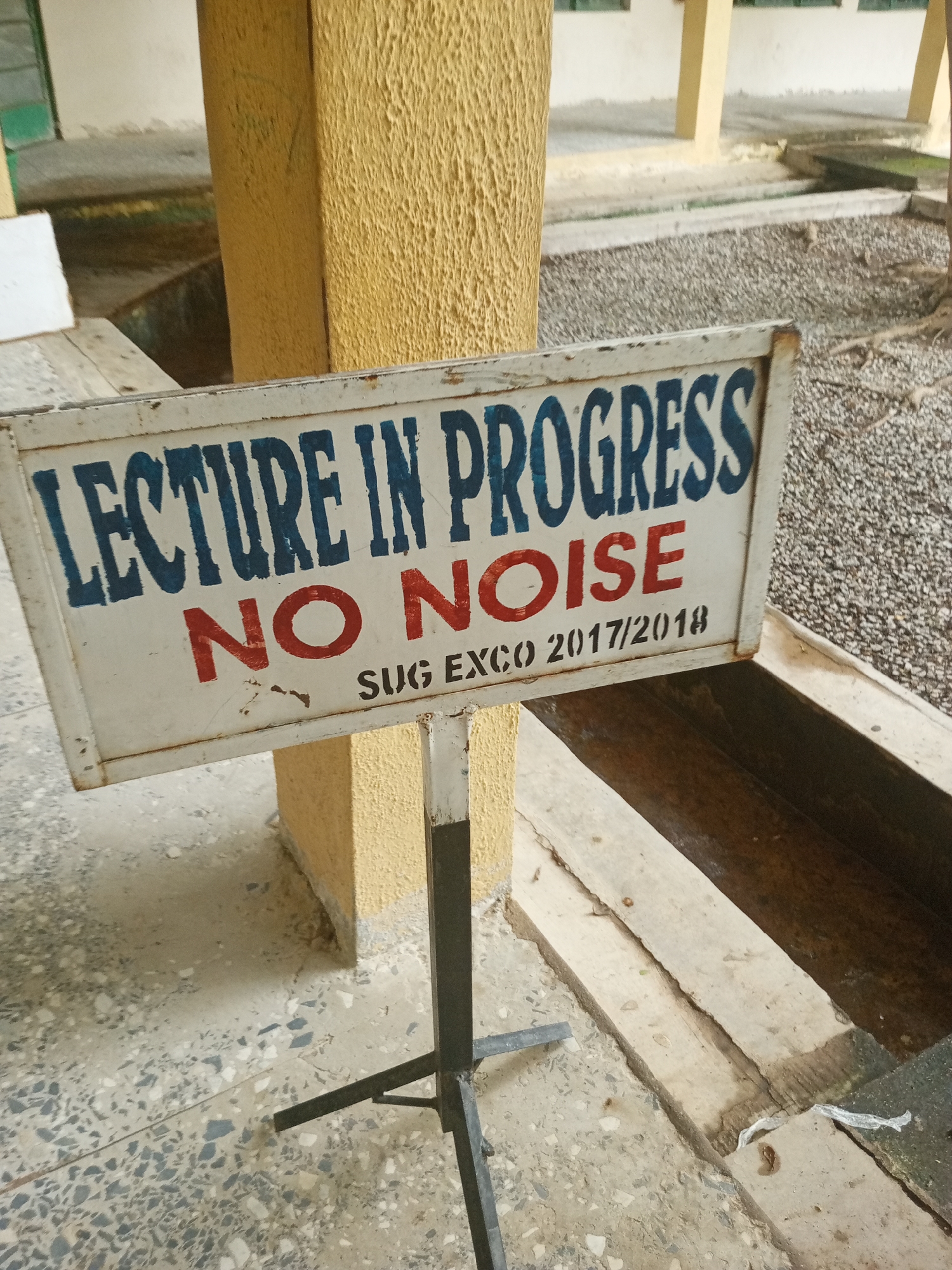 A white sign with a rustic metal frame is propped up on a black metal stand. The sign has a blue title that reads "LECTURE IN PROGRESS" in all caps. Below the title, in bold red letters, is "NO NOISE". At the bottom of the sign, in smaller black text, it says "SUG EXCO 2017/2018". The sign appears to be in an outdoor setting, possibly on a college campus or a school. There are concrete floors and gravel visible, along with some concrete structures and pillars, one of which is textured and yellow. The lighting suggests it could be daytime. The scene implies a need for quiet due to an ongoing lecture.