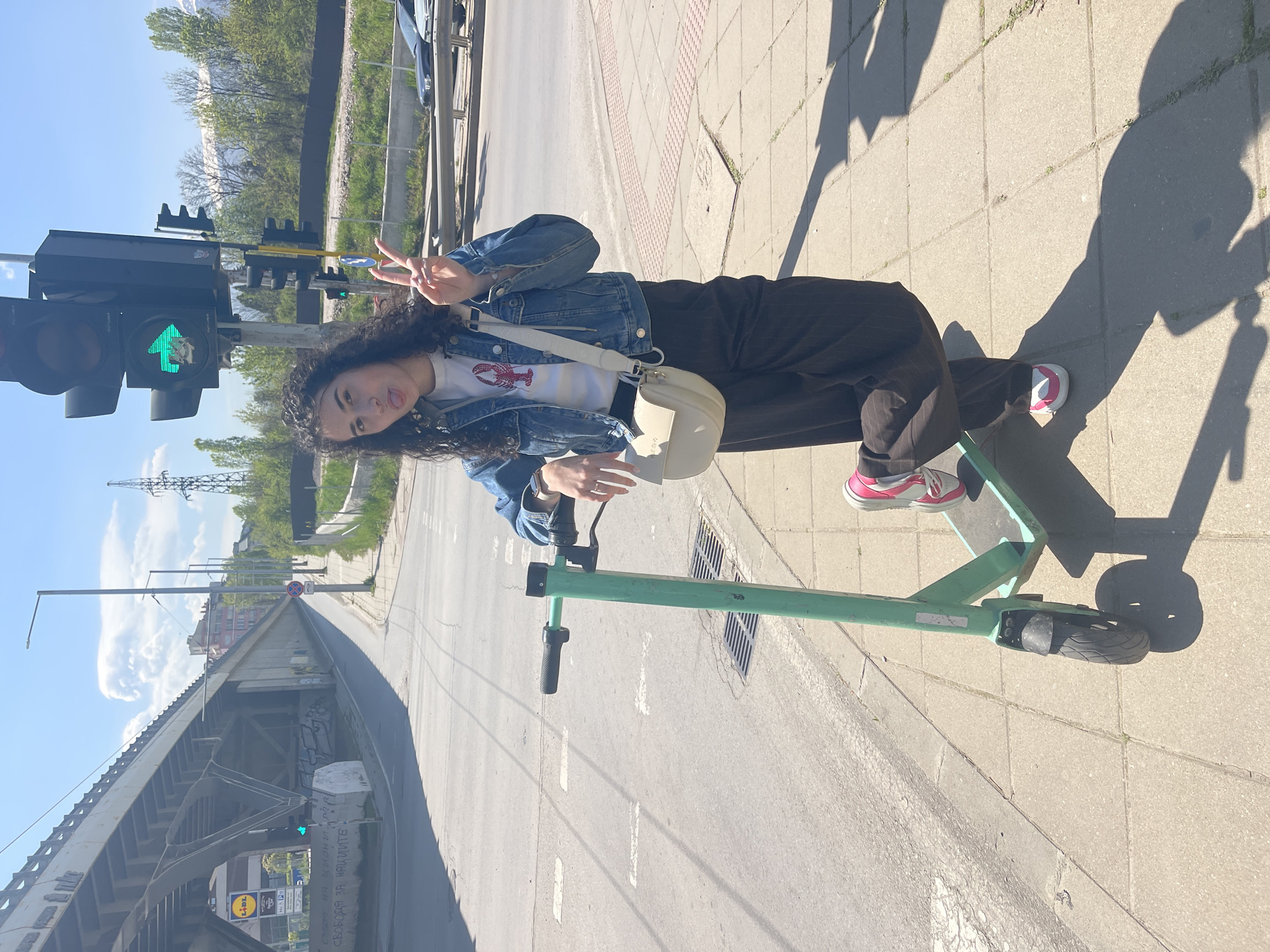 A young woman with curly dark hair is captured posing playfully on a sunny day in an urban setting, likely Sofia, Bulgaria. She stands on a paved sidewalk beside a green electric scooter, making a peace sign with her right hand and sticking out her tongue. She is dressed in a blue denim jacket over a white t-shirt adorned with a red lobster graphic, dark trousers, and white and pink sneakers. A light-colored cross-body bag, with "VALENTINO" visible on its strap, is worn over her shoulder.
The scene is well-lit by bright sunlight, casting long, distinct shadows of the woman and the scooter on the sidewalk, suggesting either morning or late afternoon. To her left, several traffic lights are visible, one prominently displaying a green arrow, indicating a clear signal for straight traffic. The background features a large concrete overpass or bridge spanning across the view. Underneath the bridge, graffiti is visible, along with a "LIDL" supermarket logo and a white sign bearing Cyrillic text, including the phrase "СВОБОДА ЗА НАРОДИТЕ" (Freedom for the People). A paved road runs alongside the sidewalk, marked with white lines. In the distance, trees, buildings, and a power line tower complete the bustling cityscape under a clear blue sky with scattered clouds.