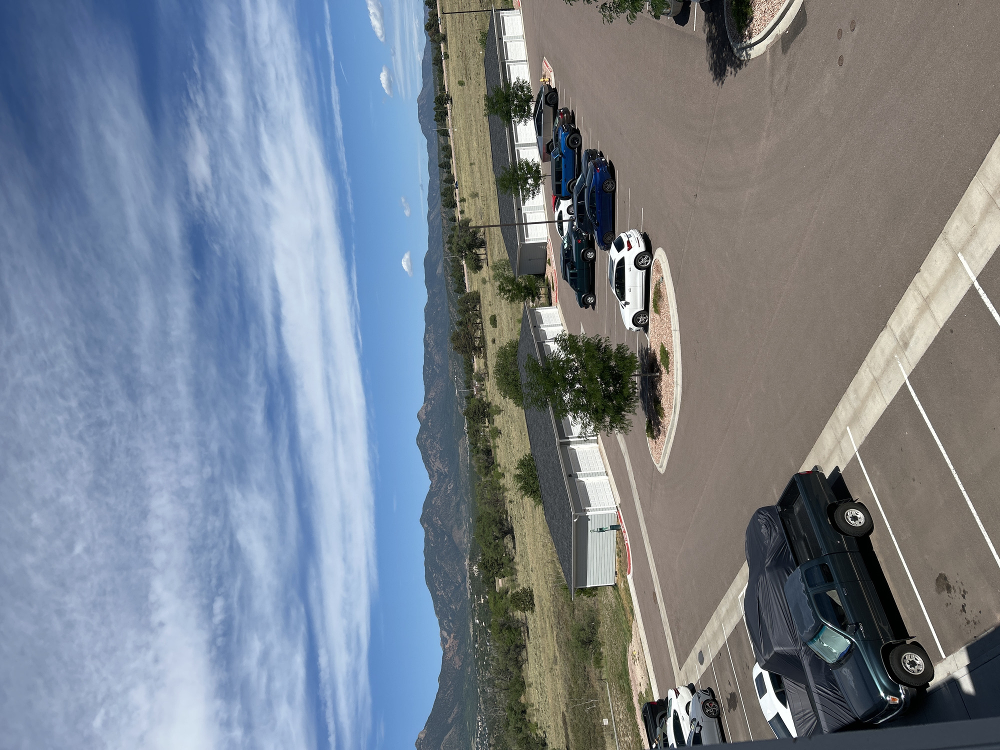 This is a high-angle, wide shot of an outdoor parking lot in Colorado Springs, USA, on a partly cloudy day. In the foreground, a dark green pickup truck is parked in a designated space. To its left, two vehicles are covered with dark tarps. Further into the lot, several other vehicles are parked, including white, blue, and gray cars and SUVs. The parking lot is asphalt with white parking lines. In the mid-ground, a row of light gray garages with white doors is visible, with small trees planted in front of them. Beyond the garages, a dry, grassy field stretches towards a range of mountains in the background. The sky is blue with scattered white, wispy clouds. The overall scene depicts a residential or storage area with a mountainous backdrop.