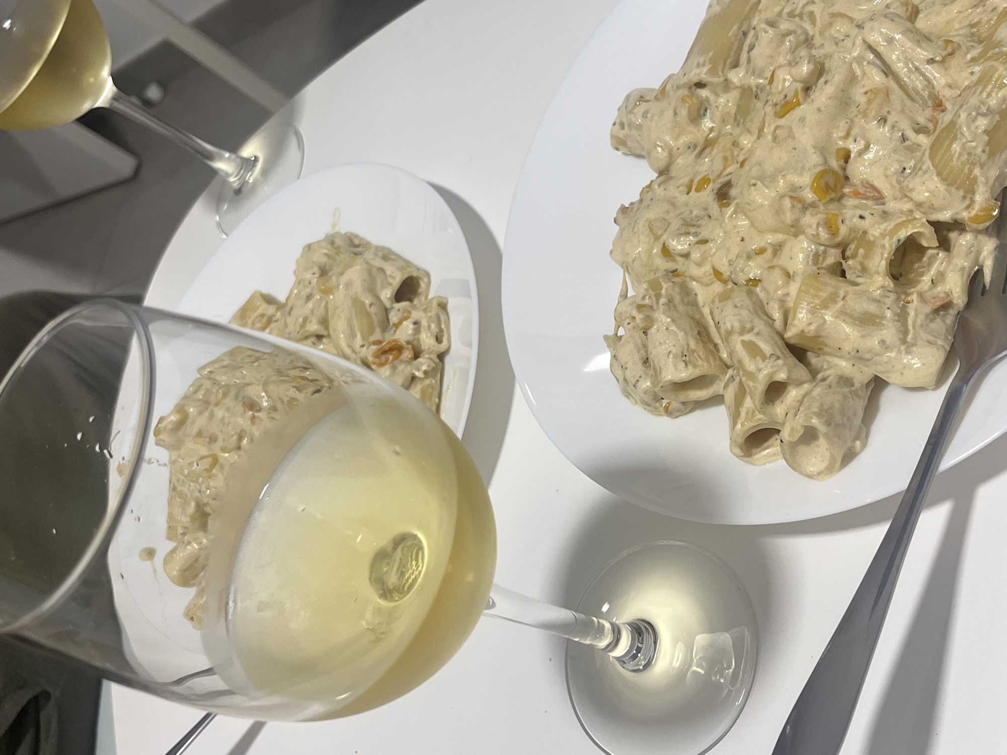 The image depicts a dinner setting for two, featuring two white plates of rigatoni pasta with a creamy sauce. A silver fork is placed on the right plate. Two wine glasses, filled with a light-colored wine, are also visible; one is in the foreground and the other is partially visible in the background. The surface appears to be white, possibly a table. The food and drink are presented on a white, likely indoor, surface. There is no evidence to suggest that this picture was taken in the city of Mogán, Spain.