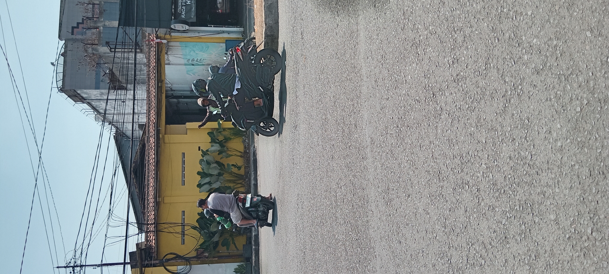 The image captures a street scene in Tasikmalaya, Indonesia, on a clear, sunny day. In the foreground, the asphalt road shows its texture of small stones. Two motorcycles are the primary subjects. On the left, a single rider wearing a grey t-shirt and light-colored shorts is on a scooter. On the right, two people are on another scooter. The rider in front is wearing a dark jacket and helmet, while the person behind them is wearing a green reflective vest and a helmet, with their right arm raised in a wave. Behind the motorcycles, a yellow building with a tiled roof and rows of dark green plants is visible. Further in the background, a taller building with a concrete facade and visible electrical wires overhead completes the urban landscape. The presence of a national flag on a pole suggests a public or commercial area.