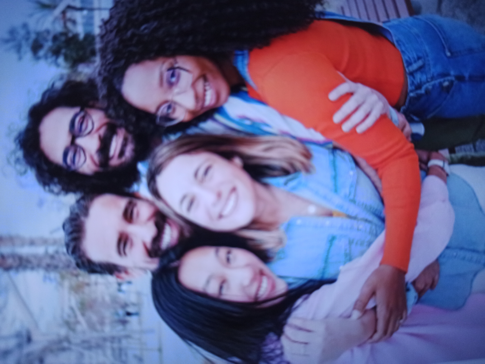In a close-up, low-resolution image, five individuals are captured in a group embrace, their faces turned towards the camera and smiling. The group consists of three women and two men. The individuals are diverse in terms of apparent ethnicity and gender. They appear to be outdoors, with blurred foliage and palm trees visible in the background, suggesting a park or an outdoor recreational area. The lighting appears to be natural daylight. The setting does not provide any specific visual cues that would indicate the location as Balakliya, Ukraine. The people are dressed casually in various colored shirts and denim overalls. The interaction is one of friendship and closeness.