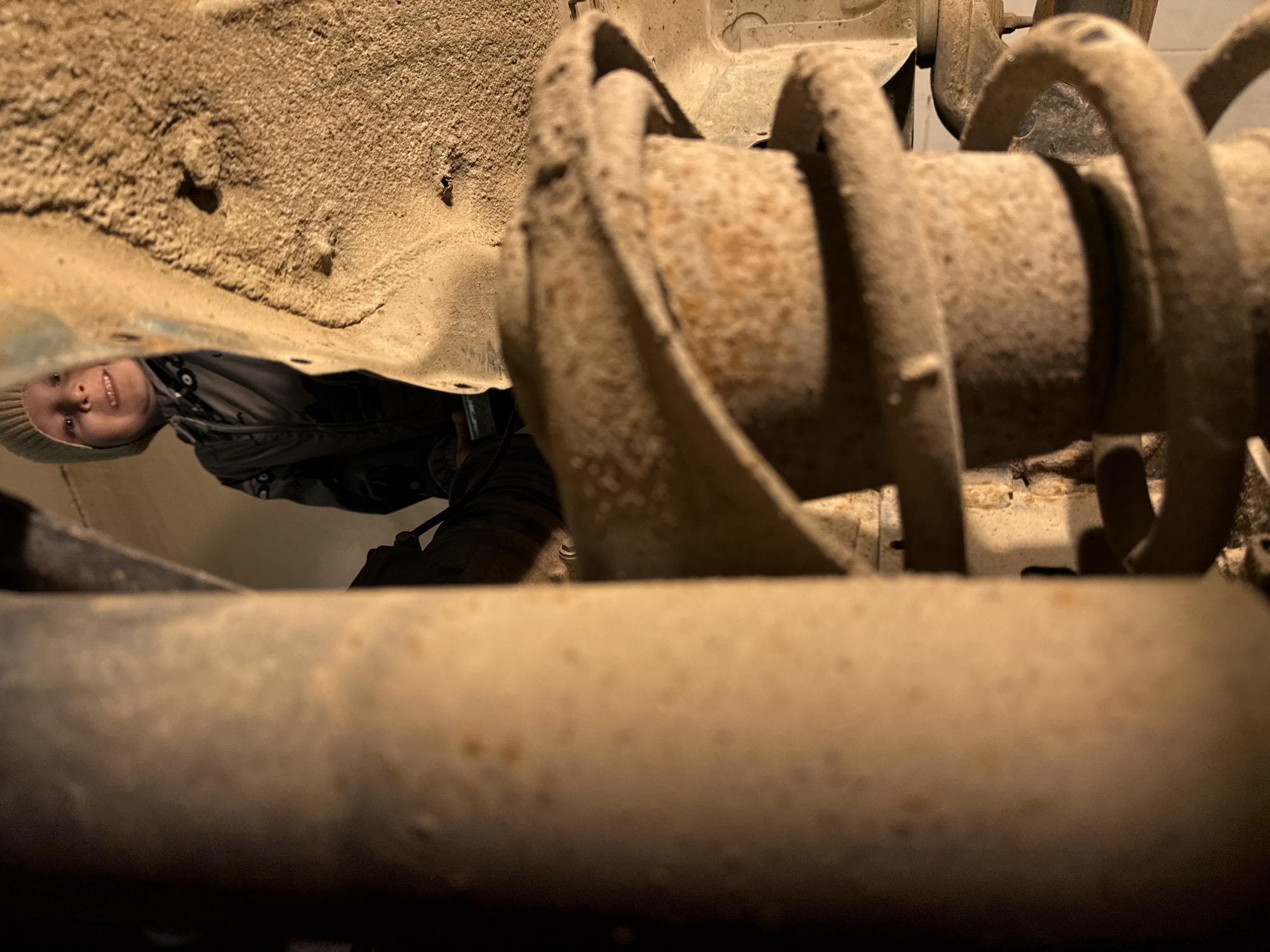 The image shows a person lying underneath a vehicle, possibly a car or truck, engaged in maintenance or repair work. The person, likely a child, is wearing a hat and jacket, looking towards the camera. Various mechanical parts of the vehicle, including the suspension coil and other undercarriage components, are visible and appear to be covered in rust or dirt. The setting appears to be outdoors, underneath the vehicle in Perm, Russia.