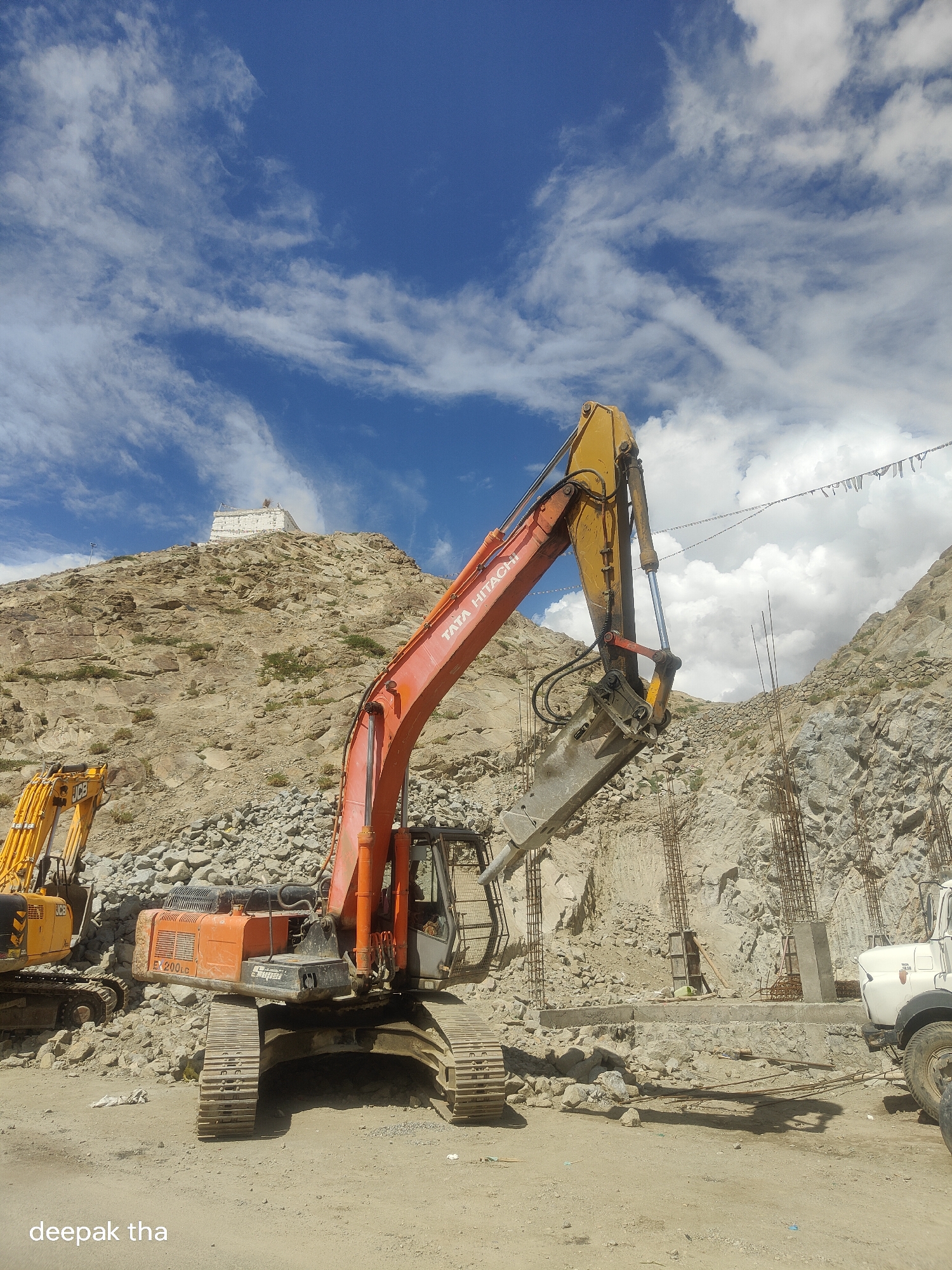 The image captures a construction scene under a bright blue sky with scattered white clouds, located in Leh, India. In the foreground, an orange Tata Hitachi excavator is prominently positioned, equipped with a large hydraulic breaker attachment, suggesting demolition or rock breaking work. To the left, the boom of a yellow JCB excavator is visible.
The background features a steep, rocky mountainside. At the summit of the mountain, a white building, possibly a monastery or a historical structure, is situated. Construction activities are evident at the base of the mountain, with rebar and concrete forms indicating the foundation of a new structure. A white truck is partially visible on the right side of the frame. The terrain is rugged and stony, typical of the mountainous landscape of Leh. The overall scene depicts ongoing infrastructure development in a challenging and scenic environment.