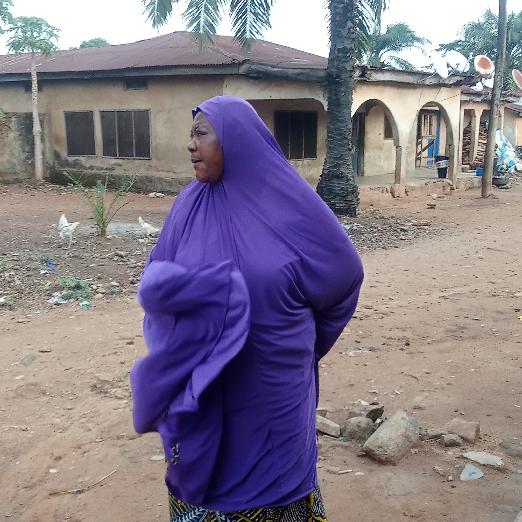 This image, taken in Akwanga, Nigeria, captures a woman standing in a semi-urban or rural outdoor setting. The woman is the central subject, positioned in the foreground and facing right in profile. She is wearing a flowing, vibrant purple hijab and a matching long-sleeved garment, with a colorful patterned skirt visible at her lower half. Her left arm is bent, and the fabric of her attire shows subtle movement.
The background features several single-story buildings with corrugated metal roofs. The structure directly behind the woman has light-colored walls and two dark, rectangular windows. To its right, another building presents distinctive arched doorways and equally light-toned walls, topped with multiple satellite dishes. Palm trees are prominent elements in the background, particularly on the right, alongside a slender tree (possibly a papaya) on the far left.
The ground is largely reddish-brown dirt, littered with scattered rocks, small pieces of debris, and some litter. Two white chickens are visible in the mid-ground to the woman's left, foraging on the dusty earth. A dark bin or barrel is noticeable near one of the arched entrances of the right-side building. The scene is illuminated by soft, diffused daylight, suggesting an overcast sky or a time of day with indirect sunlight. There is no specific event taking place, but rather a snapshot of everyday life. No visible text is present in the image.