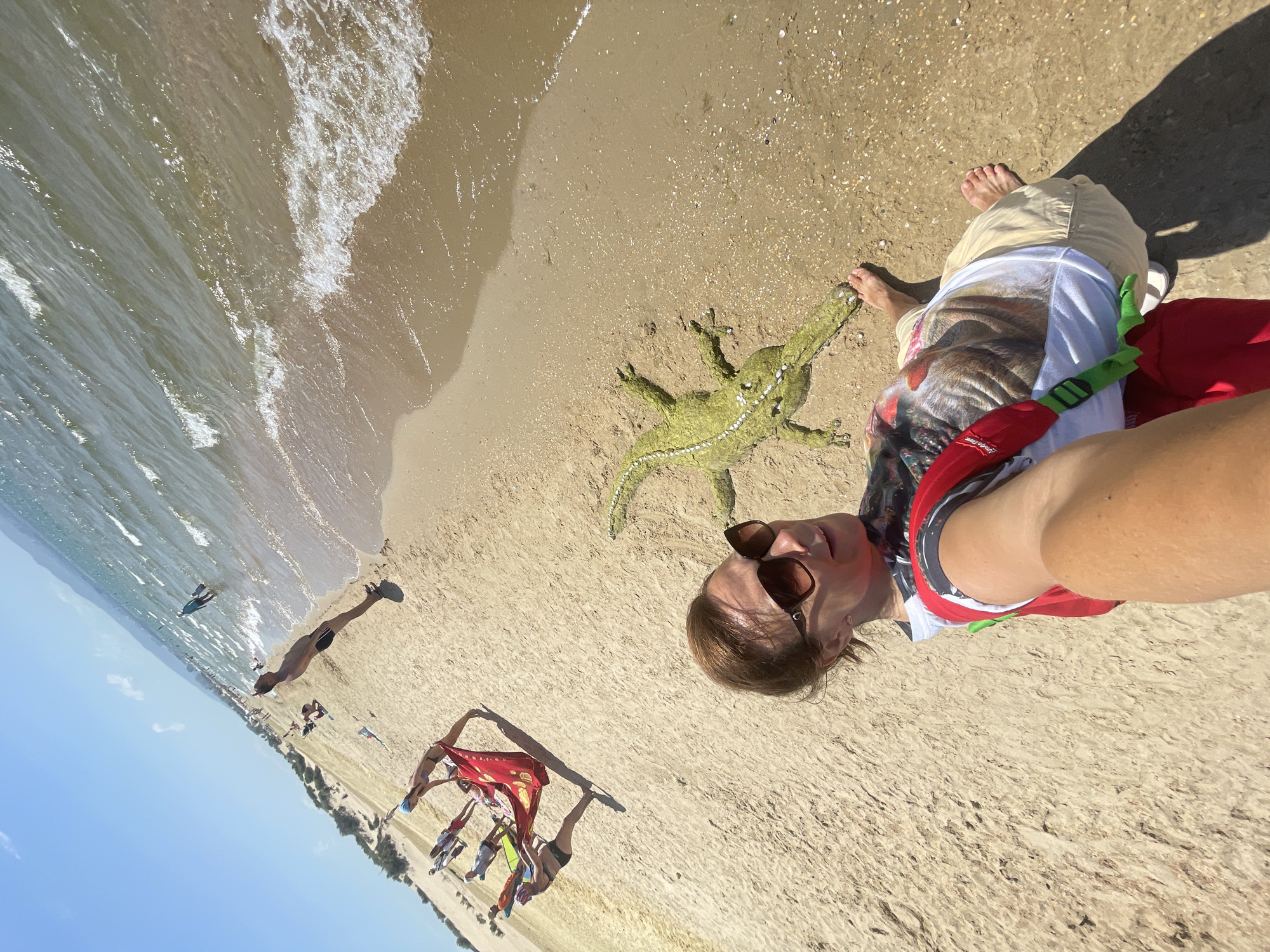 A woman is taking a selfie on a sandy beach in Anapa, Russia. She is wearing sunglasses, a t-shirt with a dog's face, and tan shorts. Next to her on the sand is a green alligator sculpture, possibly made of sand or seaweed. In the background, several people are visible on the beach and in the shallow water, some engaging in watersports like kitesurfing. The sea is calm with small waves, and the sky is clear blue. The image is captured at an angle, giving a dynamic perspective of the scene.