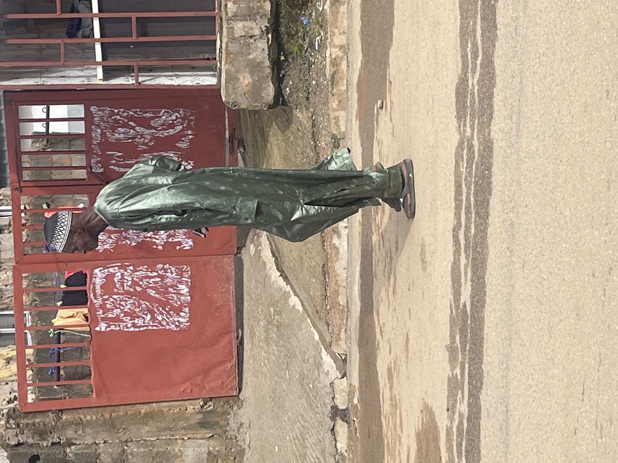 In Abuja, Nigeria, a man walks past a red metal gate. The man is wearing a long, green, shimmering robe and a white kufi cap. The gate has vertical bars and decorative white markings. Behind the gate, clothes are hanging on a line. The ground in front of the gate is made of cracked concrete and dirt, with a paved road in the foreground. The overall scene depicts a common urban street in Abuja.