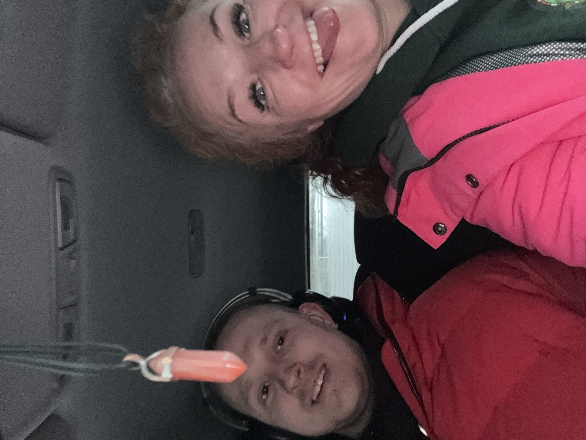 Two individuals are visible within a vehicle, with the scene appearing to be in the city of Chaikovsky, Russia.
The woman, with light skin and reddish-brown curly hair, is positioned at the top of the frame. She is wearing a pink coat over a dark green hoodie with a white stripe. Her mouth is open, tongue extended, and she is smiling at the camera.
The man, also light-skinned, is at the bottom of the frame. He has a shaved head and is wearing headphones. He is also smiling at the camera. He is wearing a red jacket over a black shirt.
In the foreground, hanging from the rearview mirror, is a small, orange cylindrical object attached to a green lanyard.
The vehicle interior is predominantly gray. The background features a partially visible building exterior through the windshield, consisting of white vertical paneling.