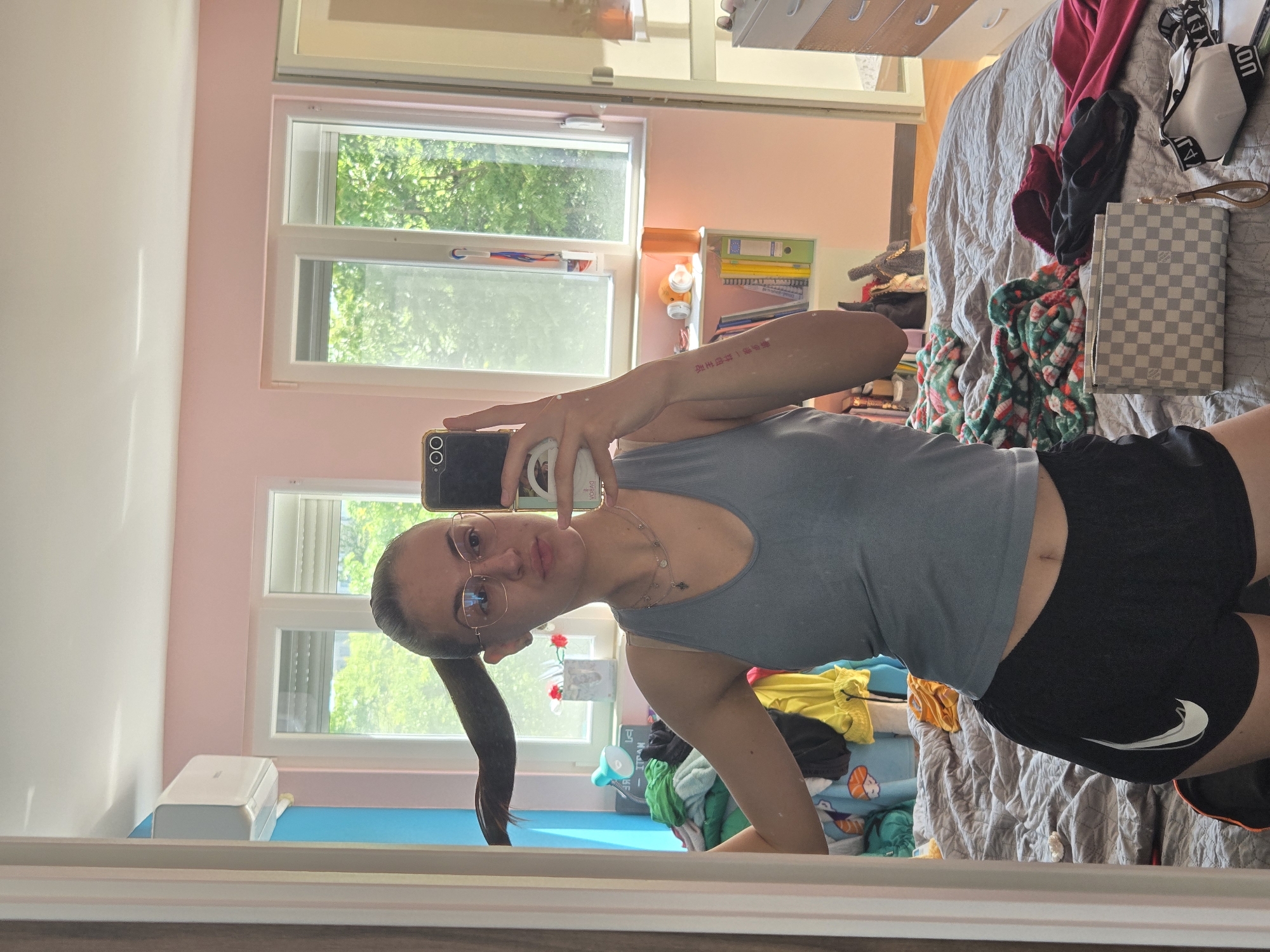 A young woman with eyeglasses and her hair in a ponytail is seen in a mirror selfie. She is wearing a gray tank top and black athletic shorts with a white Nike logo. She is holding a smartphone to take the photo. In the background, there is a bed with various items on it, including clothing, a checkered purse, and blankets. A window provides natural light, and there are visible trees outside. Additionally, a white air conditioning unit is seen below the window. The photograph appears to be taken indoors in a bedroom setting. The city of Pleven, Bulgaria, is not discernible from the provided image.