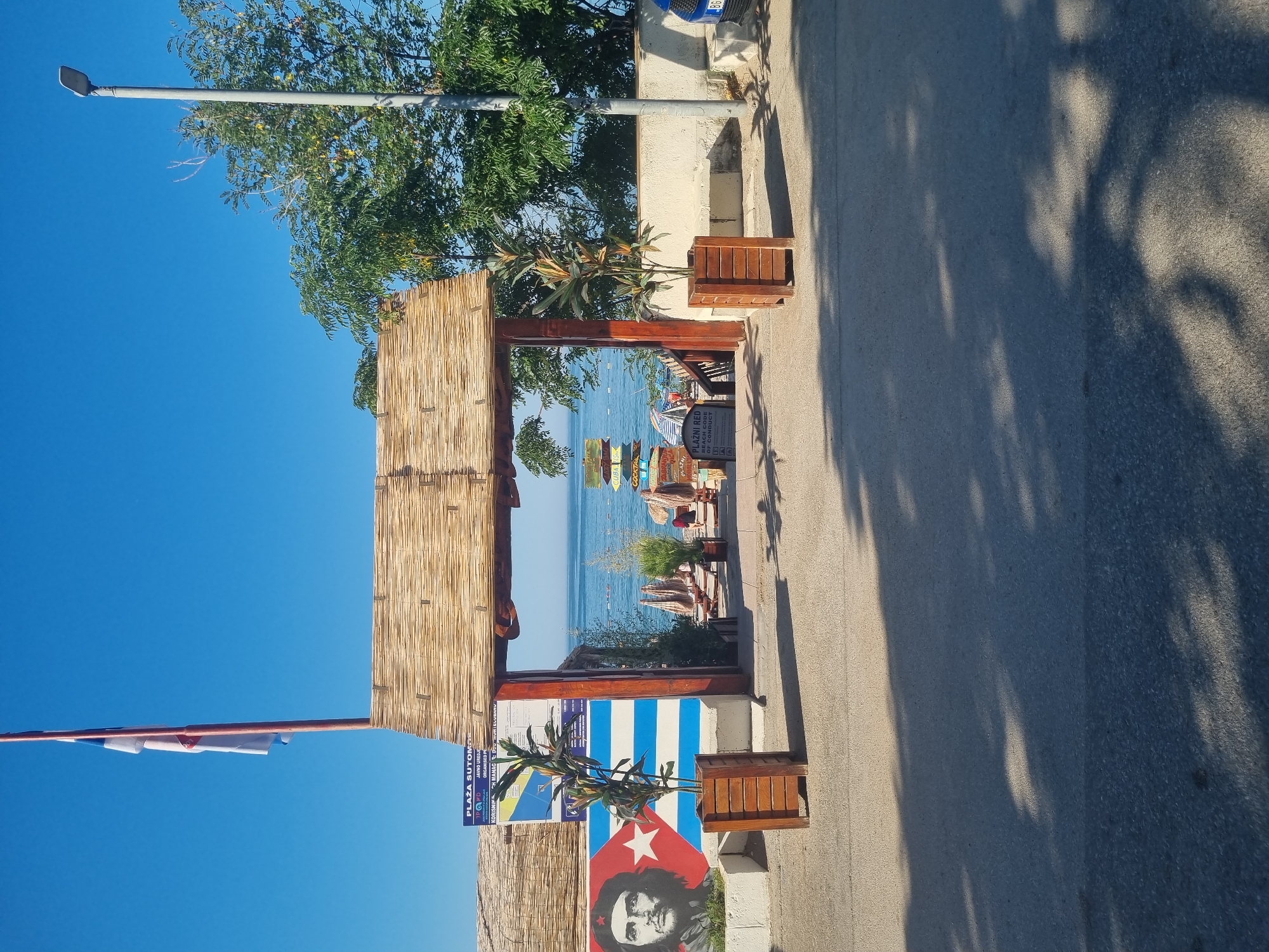 The image captures an entrance to a beach in Sutomore, Montenegro, characterized by a rustic, thatched-roof archway. To the left of the arch, a mural of Che Guevara is painted on a wall, superimposed on the Cuban flag's stripes. Two large wooden planters flank the entrance, each containing a young plant. Beyond the arch, the beach is visible with rows of lounge chairs and umbrellas, and the calm sea stretches to the horizon under a clear blue sky. A flag flies on a tall pole above the archway. A street lamp stands to the right of the scene, and shadows of trees are cast on the asphalt road in the foreground.