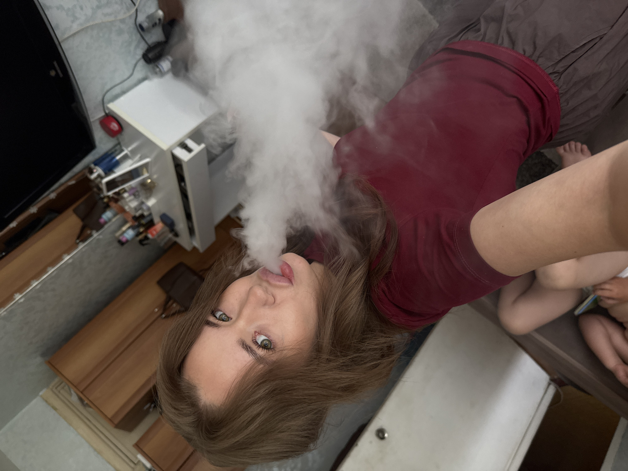 A young woman with brown hair and green eyes exhales a cloud of smoke. She is wearing a maroon t-shirt and grey shorts. The image appears to be taken indoors, possibly in a bedroom or living area, with wooden furniture and a white dresser visible in the background. The location is described as Chaikovsky, Russia, though there are no specific visual cues in the image to confirm this.