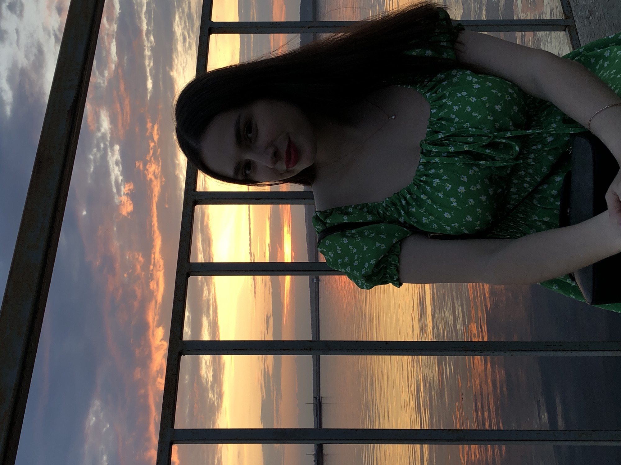 A woman with dark hair is standing on a bridge or overlook in Chaikovsky, Russia. She is wearing a green floral dress. The sky is a mix of blue, orange, and yellow colors indicating a sunset or sunrise. The setting is reflected in the water below. A metal railing surrounds the area where she is standing.
