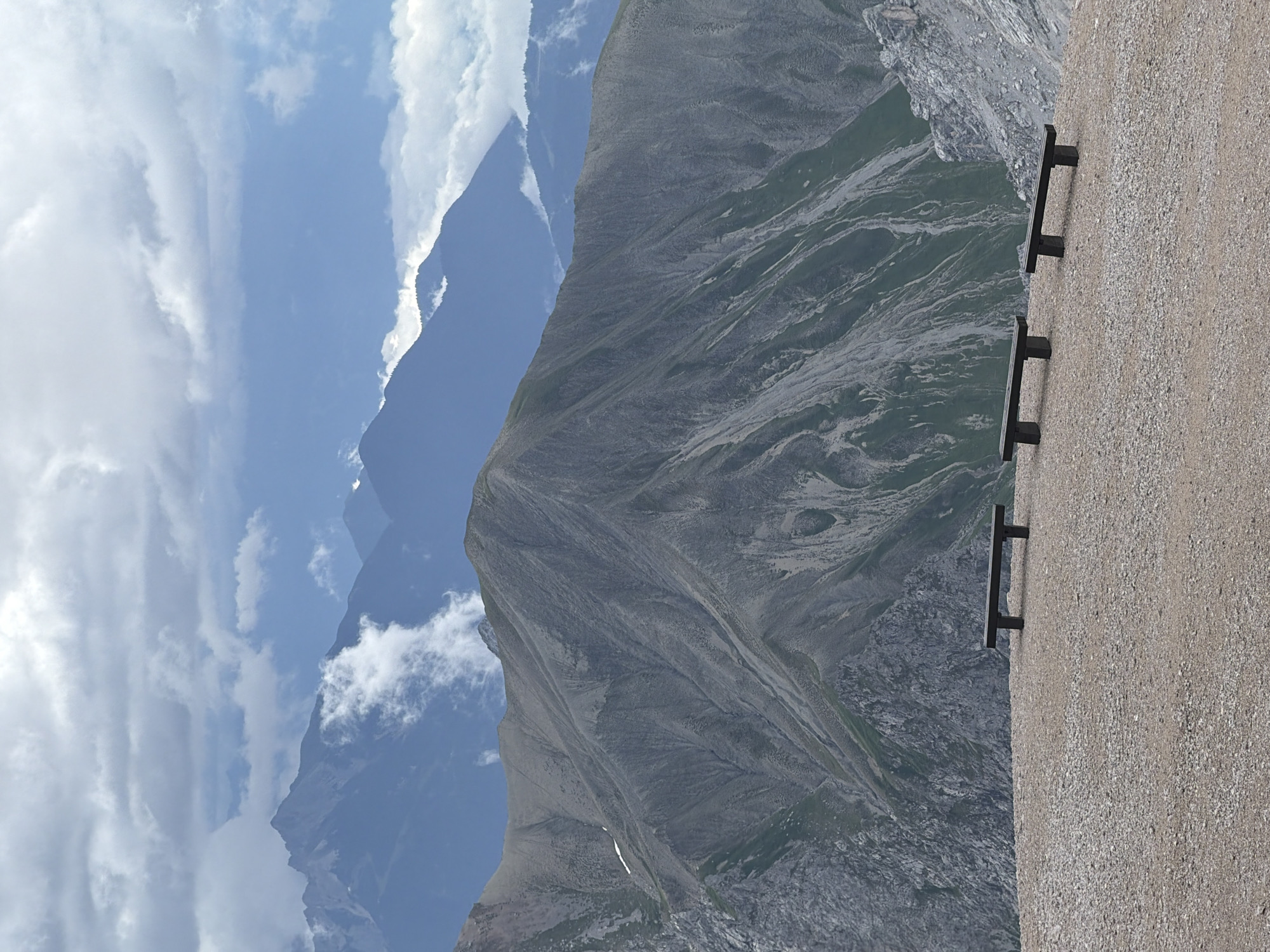 The image depicts a panoramic mountain landscape with three dark, simple benches positioned on a gravelly plateau in the foreground. Behind the plateau, a large, rugged mountain dominates the mid-ground, its slopes showing a mix of grey rock and green vegetation. In the background, layers of further mountain ranges recede into the distance, partially obscured by clouds and a hazy blue sky. The scene suggests a location at a high altitude, likely a viewpoint or hiking trail, offering expansive views of the mountainous terrain.