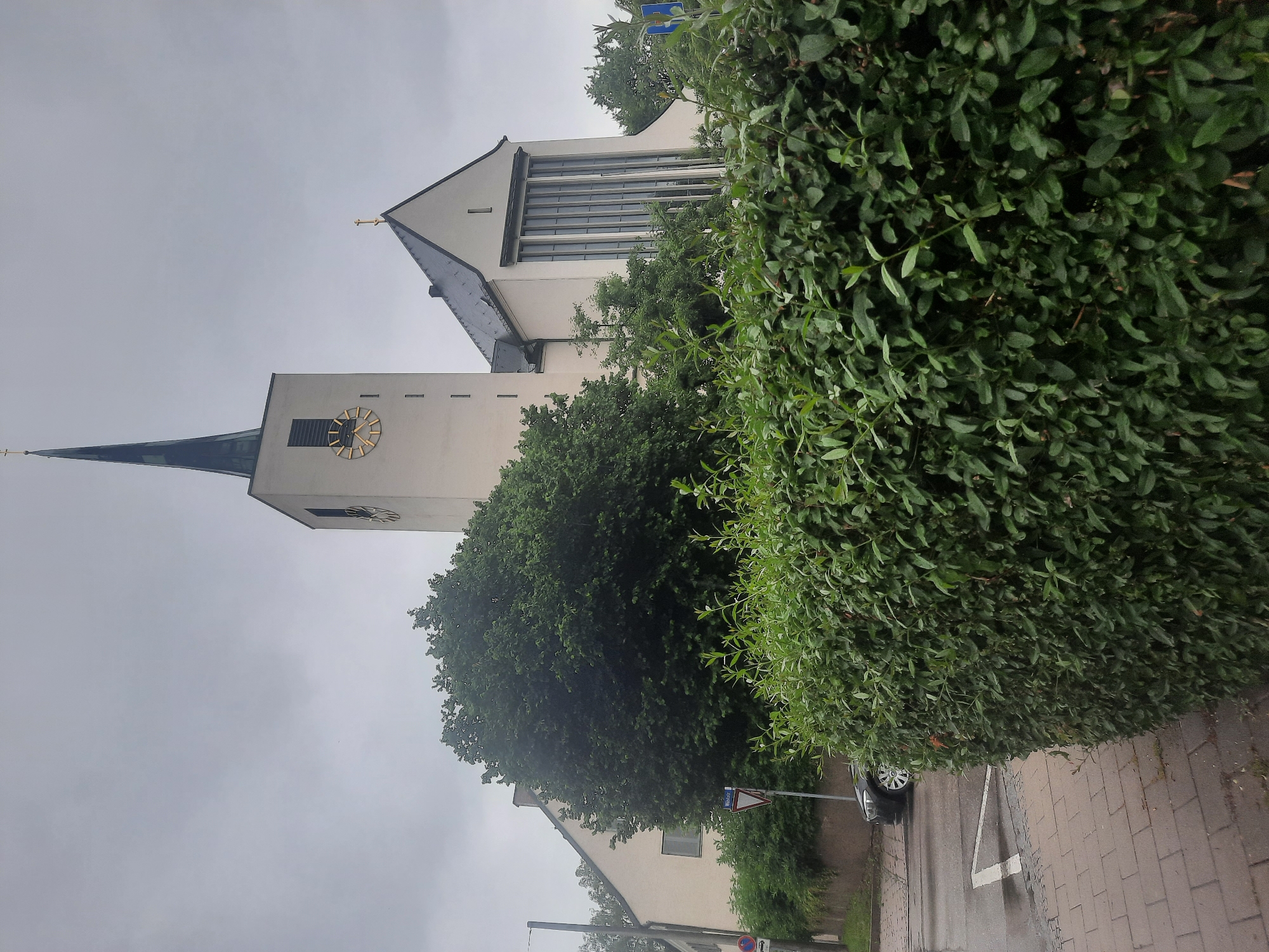 The image depicts the exterior of a church in the city of Rosenheim, Germany. The church features a tall, slender steeple with a dark roof, topped by a golden cross. A clock face is visible on the steeple. The main building of the church is white with a dark roof and features large, vertical windows. In the foreground, a dense, well-maintained green hedge runs across the frame. To the left of the church, a house with a light-colored facade and a dark roof is visible, partially obscured by a large, full tree. A street runs in front of the house and church, with a black car parked on the wet asphalt. A brick sidewalk is visible in the lower left corner, with a white line marking what appears to be a crosswalk. A street sign is mounted on a pole near the house. The sky is overcast and grey, suggesting a cloudy or rainy day.