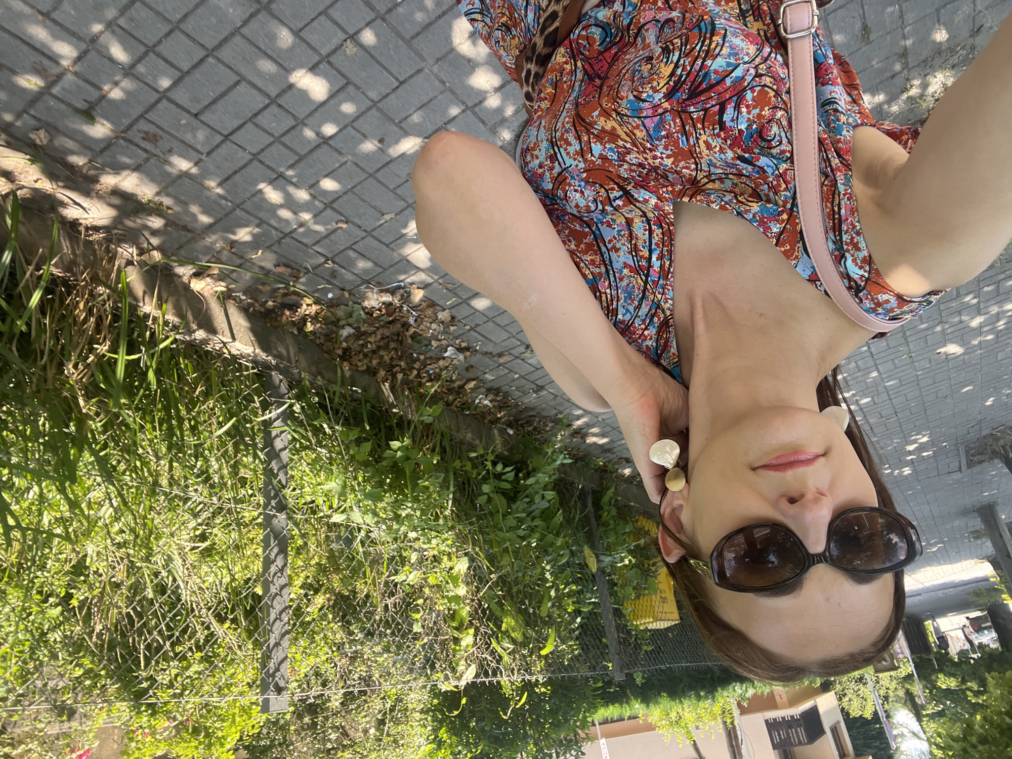The media file is a selfie taken outdoors, likely on a sunny day, judging by the dappled light on the pavement. The subject is a woman wearing a brightly colored, patterned dress and sunglasses. She is also wearing large, circular earrings. A pink purse strap is visible over her shoulder. The background includes a paved area with square tiles, grass, and a chain-link fence. There are trees and buildings visible in the distance, suggesting an urban or suburban environment. The woman is looking towards the camera, and her expression appears neutral. The image is rotated 90 degrees clockwise.