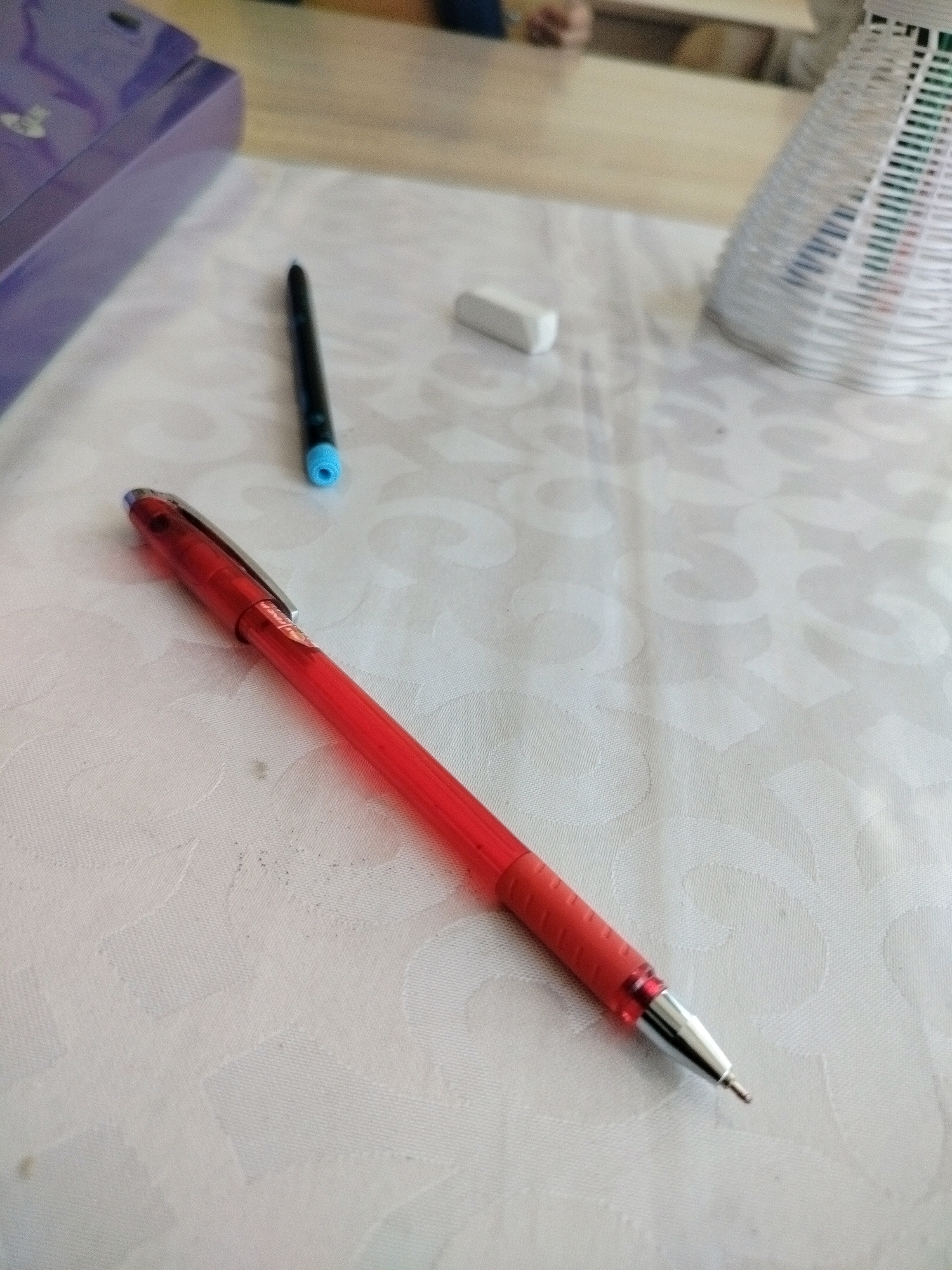 A red pen lies on a white tablecloth with a subtle pattern, angled diagonally. Behind it, a black pen with a blue tip stands upright. Further back, a small white eraser is visible. To the left, a portion of a purple object, possibly a box or container, is in the background, partially out of focus. To the right, a white woven basket with items inside is also present. The scene appears to be indoors, possibly on a table. There are no people clearly visible, but a blurred figure can be seen in the far background. The location is specified as Көлкент, Kazakhstan.