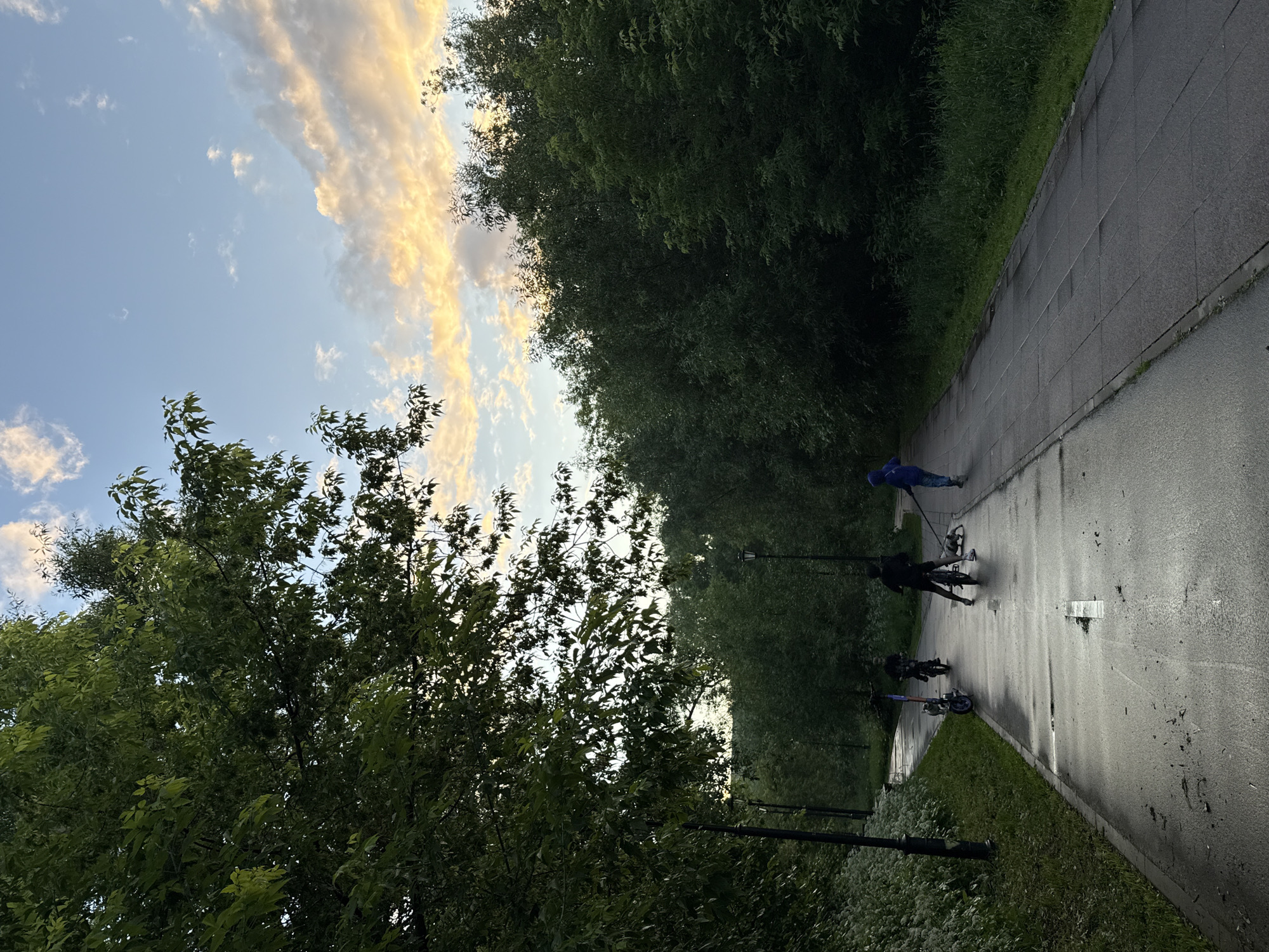 The image depicts a paved path in a park, likely in Moscow, Russia, given the presence of birch trees, common in Russian parks. The path is wet, suggesting recent rain, and is lined with lush green trees and vegetation. Several people are present: two individuals on electric scooters, one person on a bicycle, and another individual walking a dog. The sky above is a mix of blue and clouds illuminated by the warm hues of either sunrise or sunset. Lampposts are visible along the path. The overall atmosphere is serene, with people enjoying outdoor activities in a natural setting.