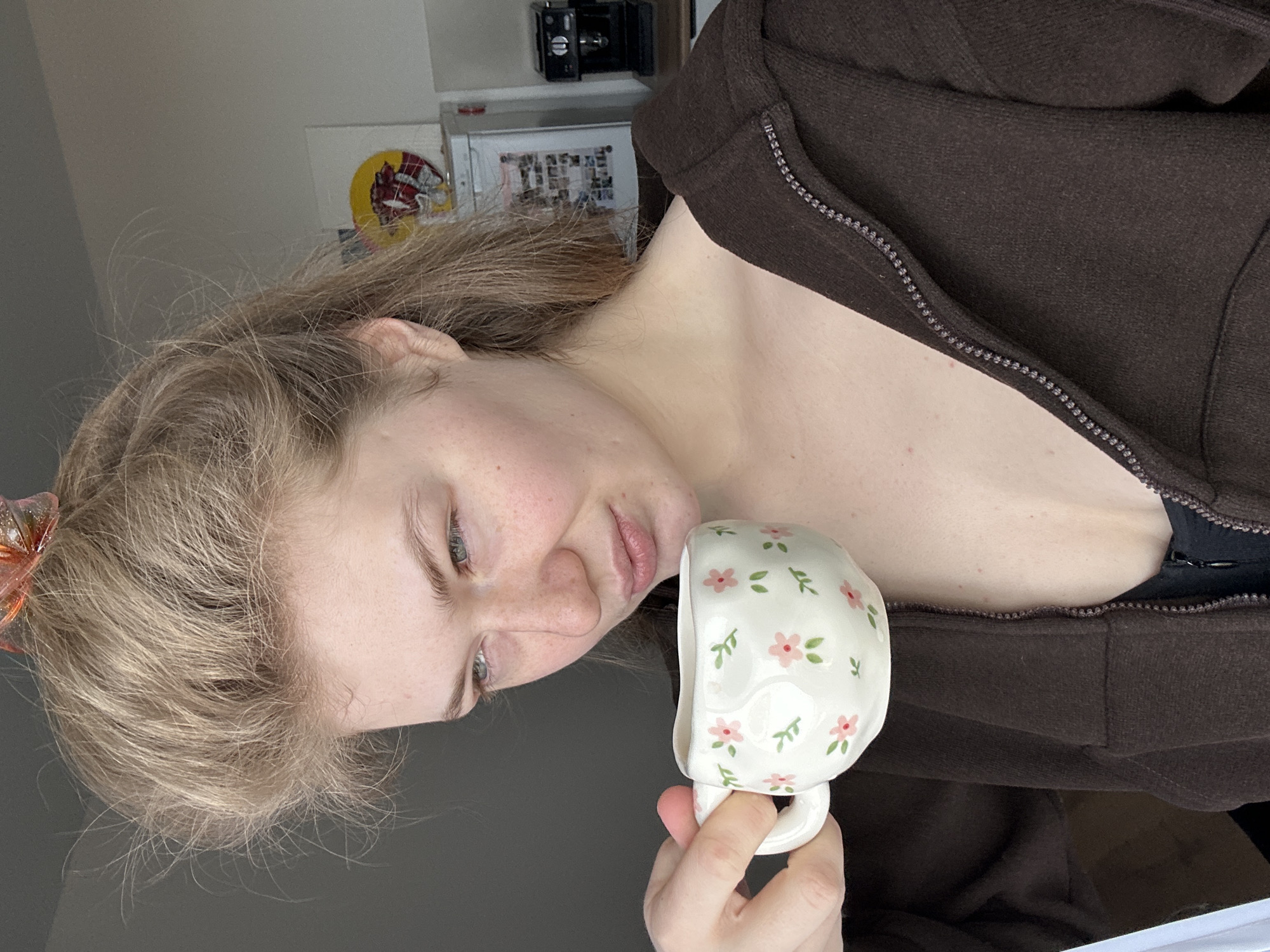 A young woman with fair skin and blonde hair holds a white ceramic mug with a floral pattern. She wears a brown hooded zip-up sweatshirt over a black top. The background includes a white shelf with various items, including a black camera, photos, and a circular object with a red and yellow design. The location is not discernible from the image; there is no indication of Kazan, Russia.