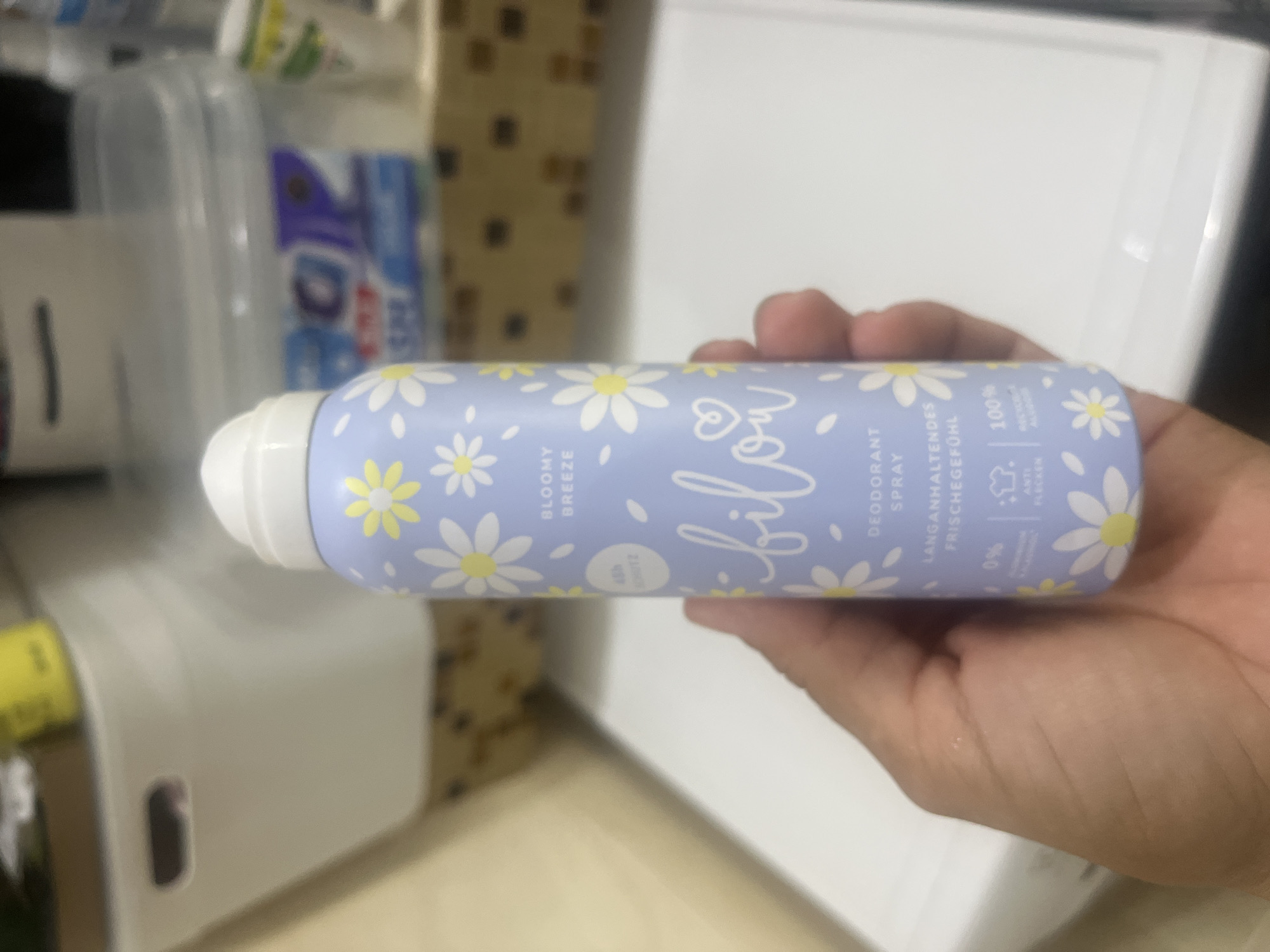 A person's hand holds a light purple aerosol spray can adorned with white daisies. The can has white text that reads "bilou" in a stylized font, with a heart symbol above the "o". Above "bilou" it says "BLOOMY BREEZE" and below it "DEODORANT SPRAY". The can also has text in German that translates to "long-lasting freshness". The background appears to be indoors, with white and tan surfaces visible. There are also other blurred objects in the background, possibly containers or other household items. The specific location of Onokivtsi, Ukraine is not discernible from the image.