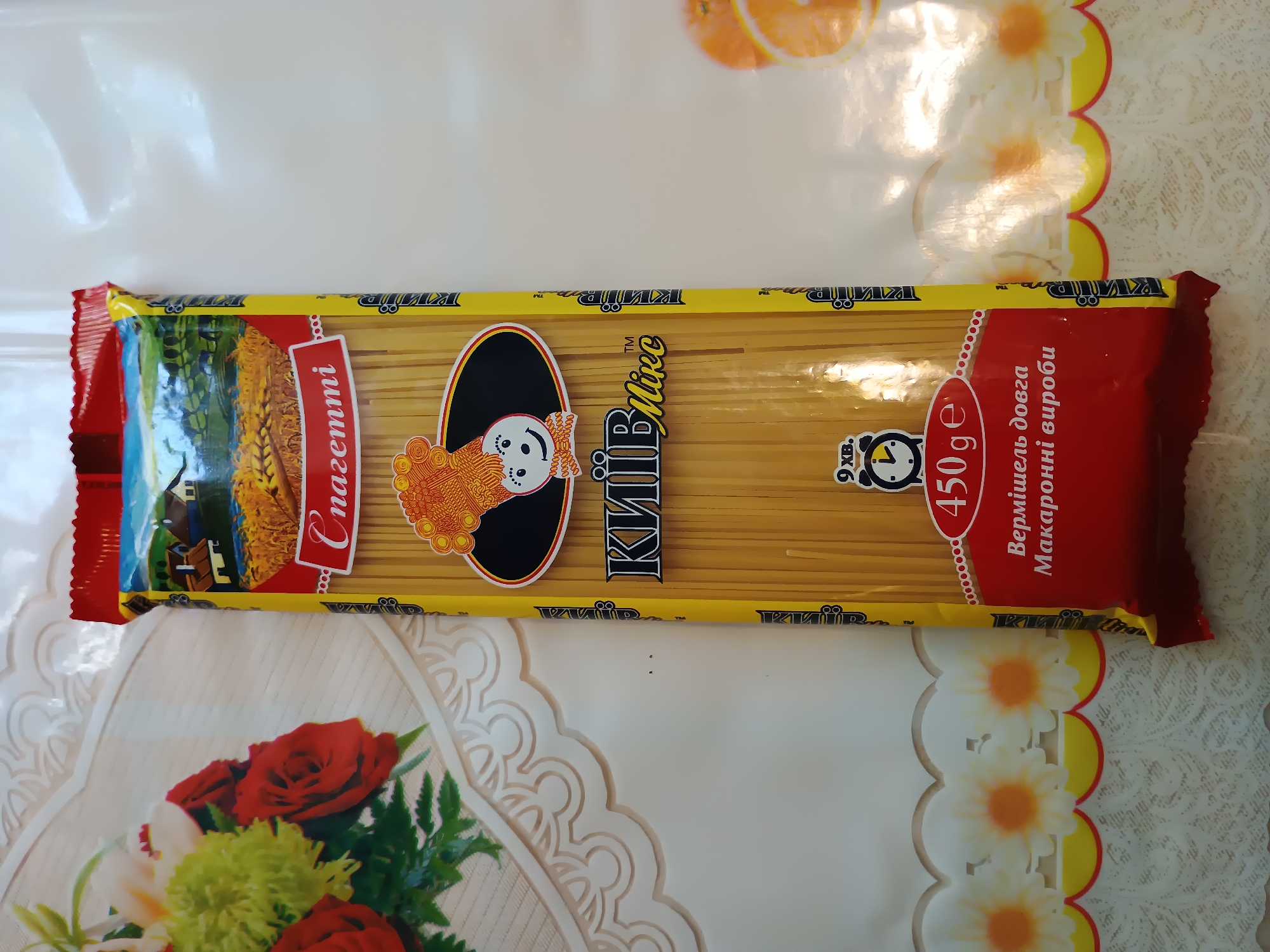 The image shows a package of spaghetti positioned vertically on a white tablecloth with a floral pattern. The spaghetti package is yellow with red accents and features a cartoon chef character and text in a foreign language. The background is out of focus but appears to be a room with ambient lighting. There are no people or animals visible in the image. The image does not contain any information or visual cues that would allow for the identification of a specific location in the city of Zaporizhzhia, Ukraine.