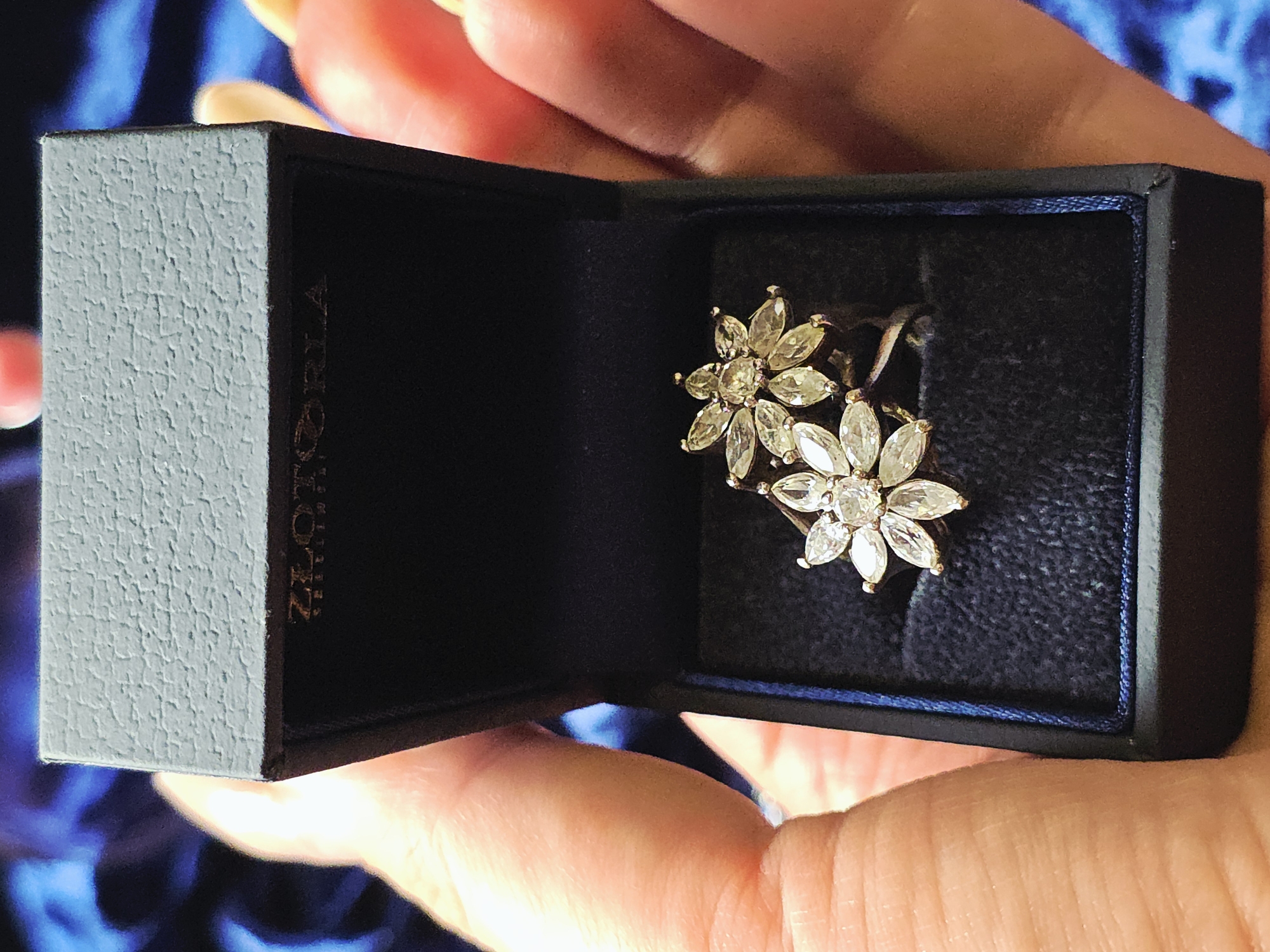 A pair of flower-shaped earrings with marquise-cut stones are displayed in an open, dark blue jewelry box held in a person's hands. The box has the word "ZLATORIA" vertically printed on its side. The backdrop is blurred, with blue tones suggesting fabric. The location is Unknown, Unknown.